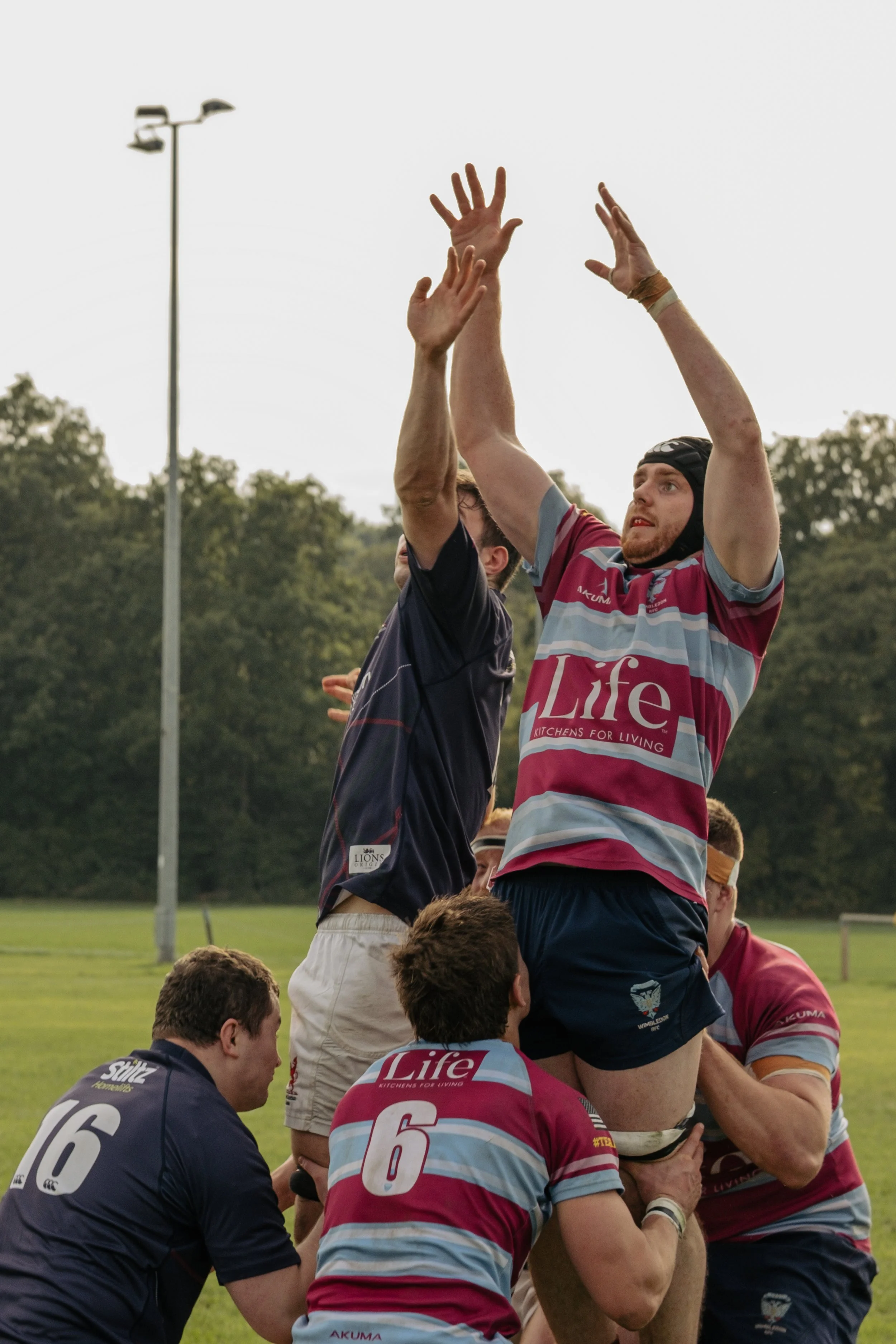 Rugby players in a lineout, with one player being lifted by teammates to catch the ball during a match on a grassy field, with trees and a lamp post in the background.