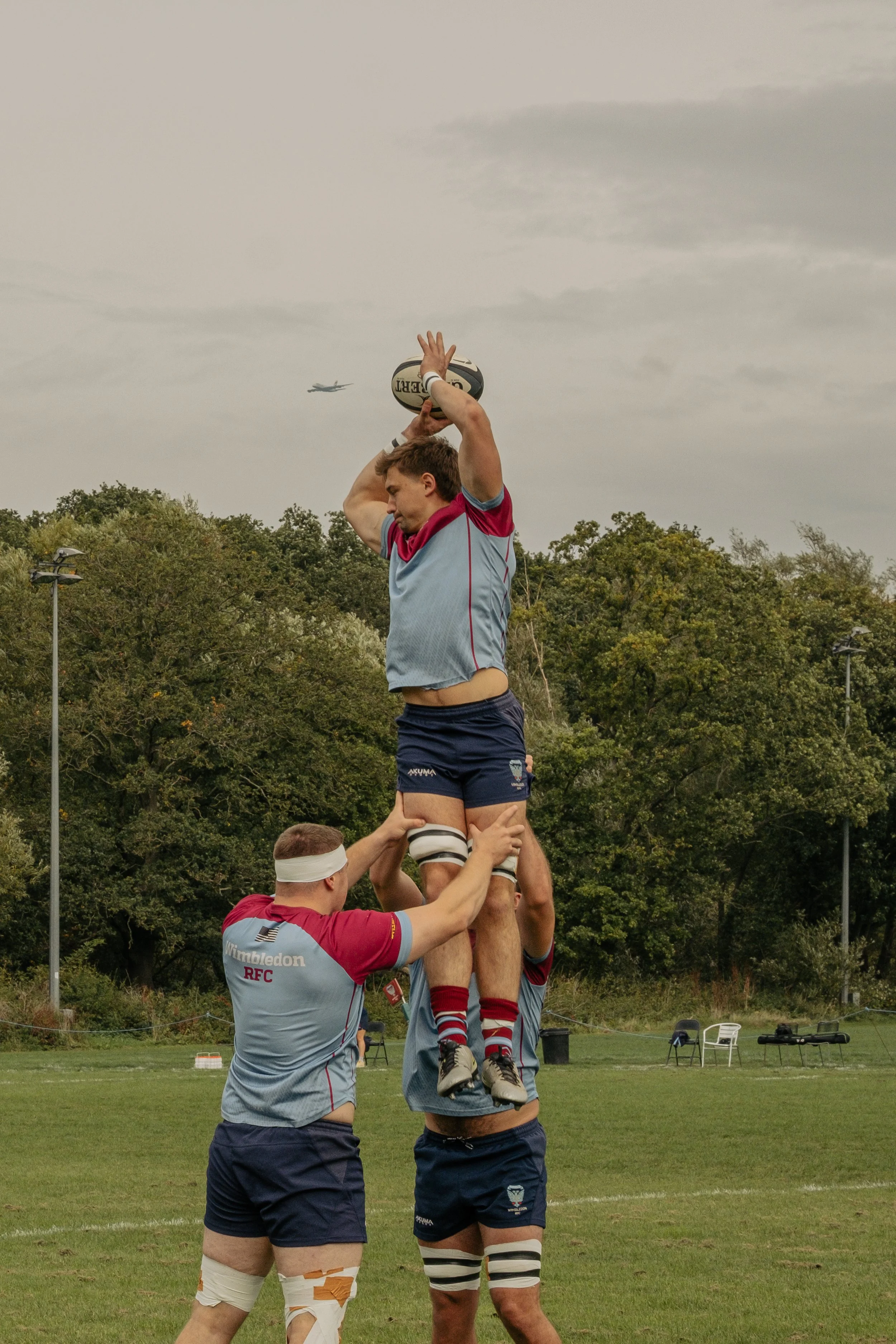Rugby players performing a line-out on a rugby field with trees and cloudy sky in the background.