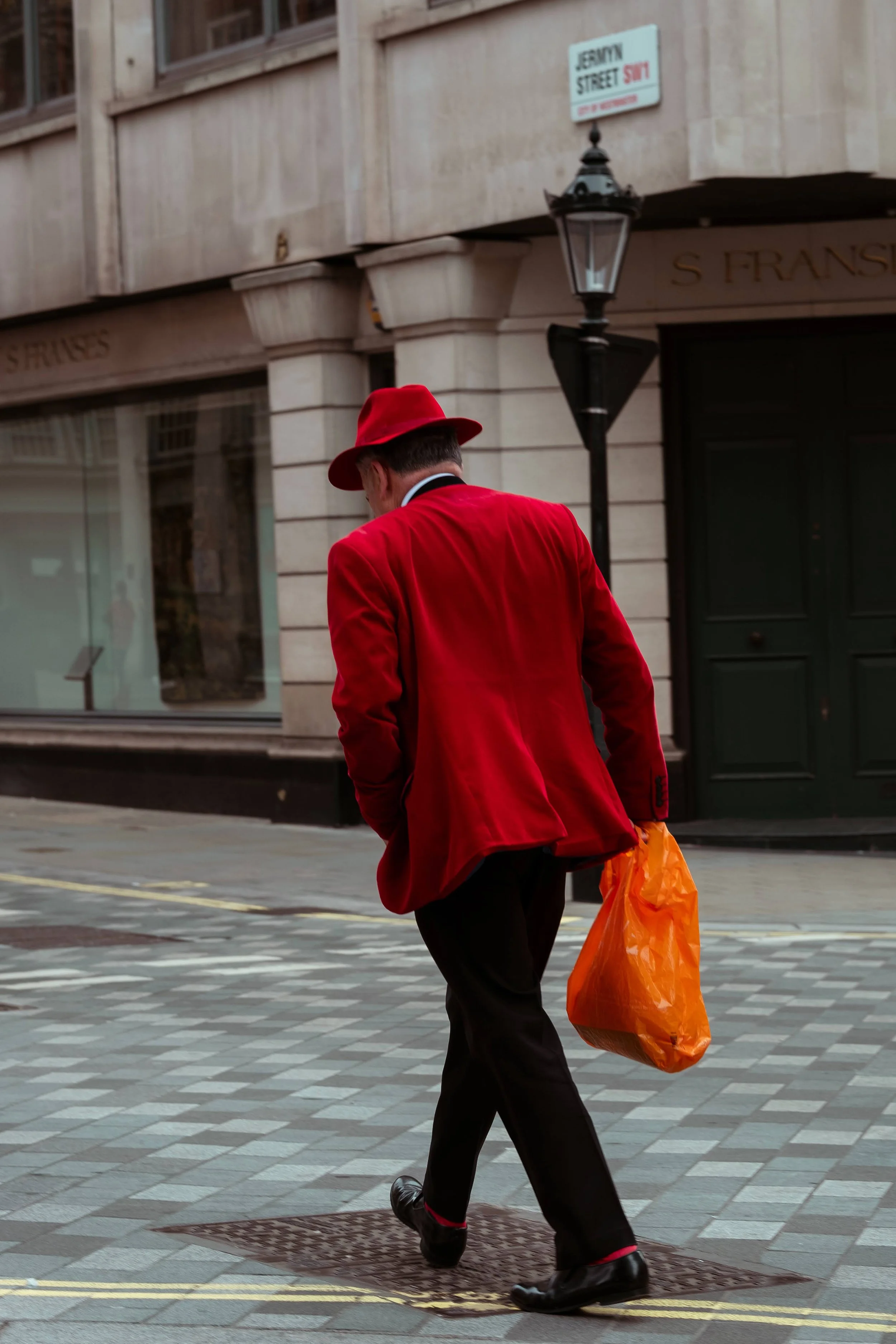 A man walking on a city sidewalk wearing a red blazer, red hat, and black pants, carrying an orange shopping bag.