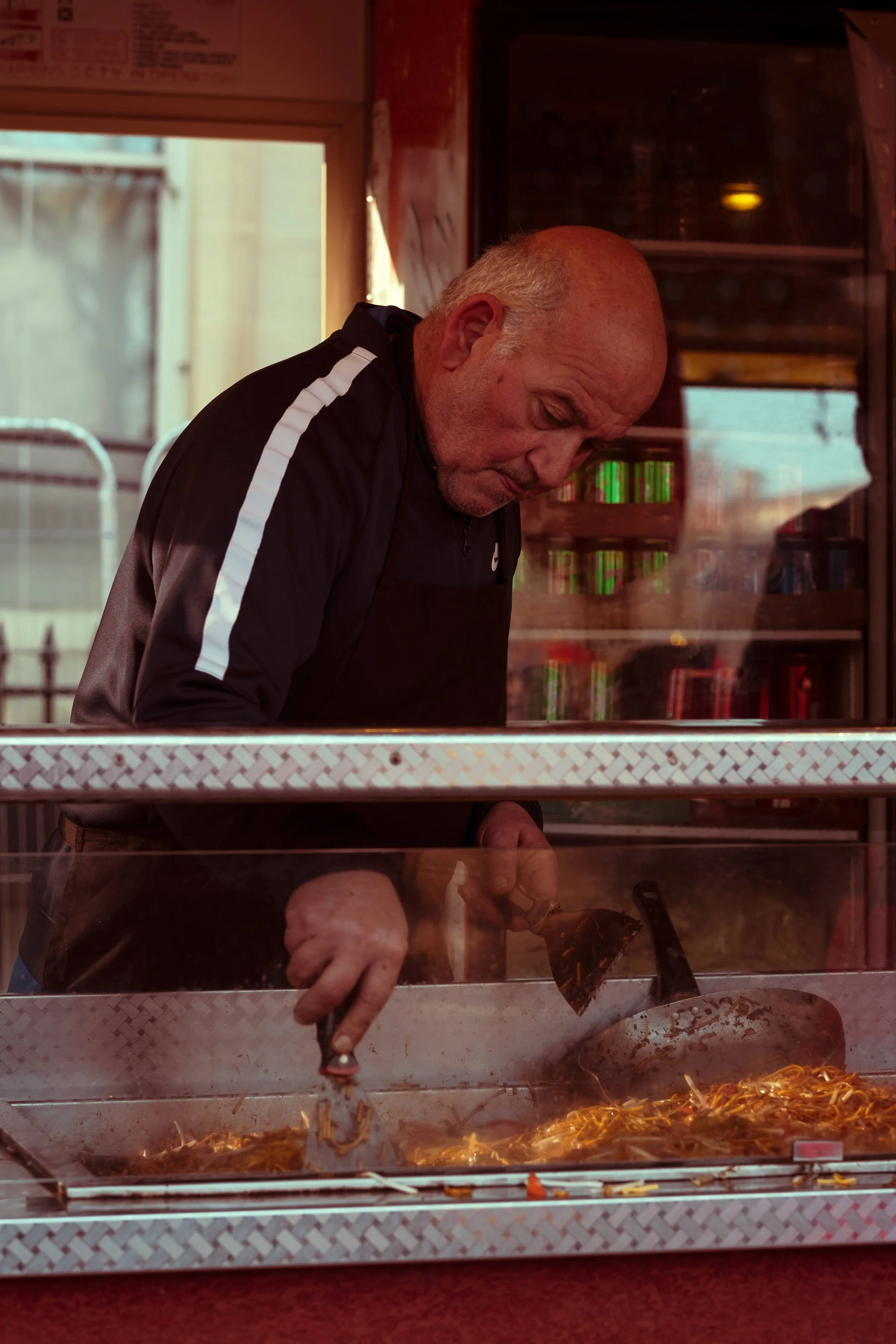 An elderly man with a bald head cooking noodles on a food stall counter, looking down at the cooking process.