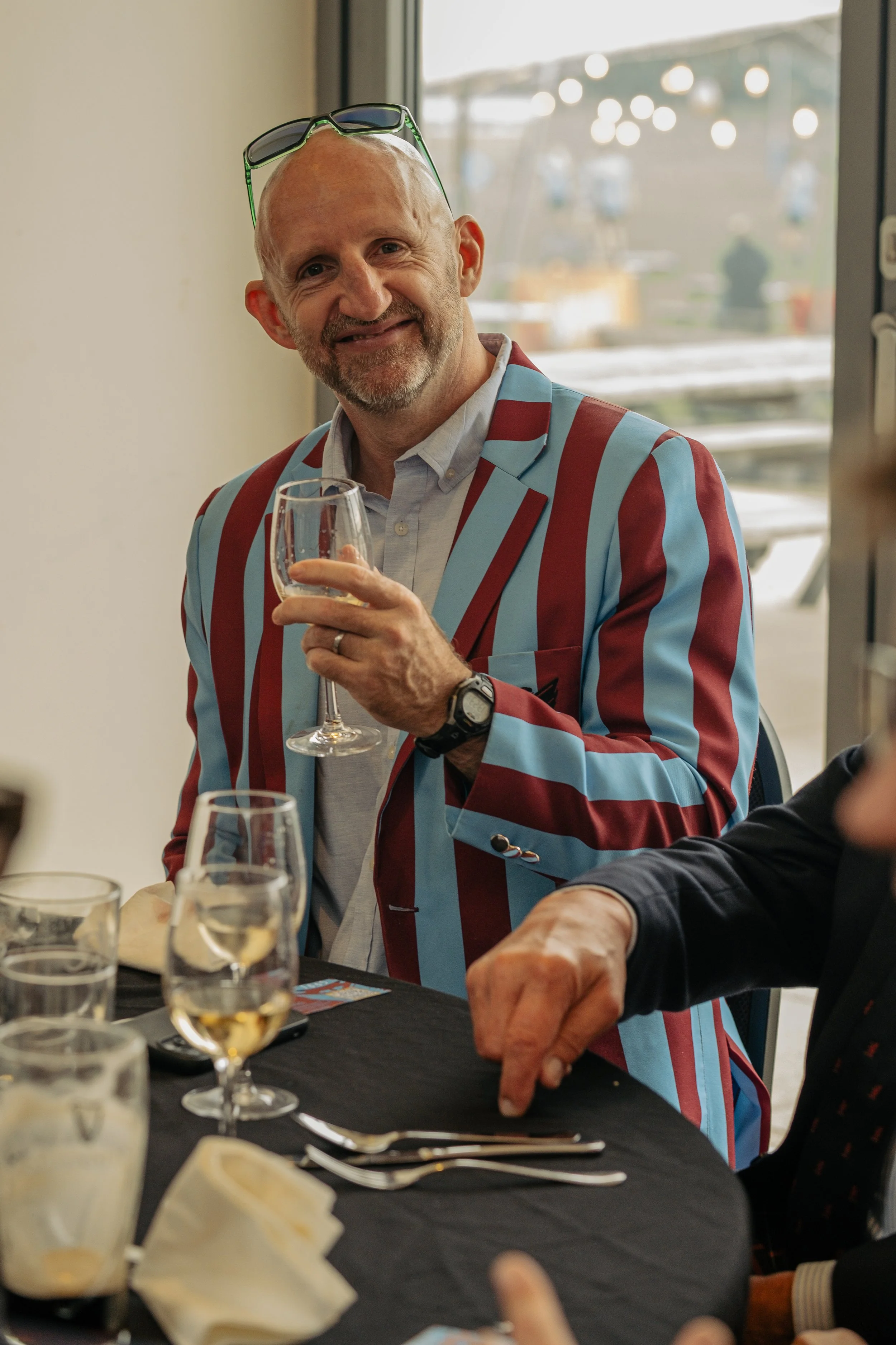 A man with a bald head, beard, and glasses on his forehead, wearing a blue and red striped blazer, holding a glass of white wine, smiling at the camera, seated at a table with glasses and utensils, with a window in the background.