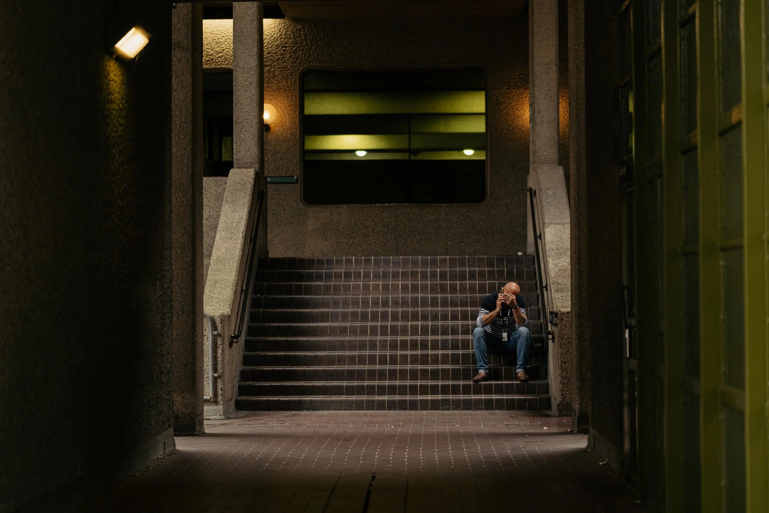 A man sitting on stairs in an underpass, holding a camera to his face, positioned in the lower right corner of the image. The stairs lead up to an underground walkway. The surroundings are dimly lit with wall-mounted lights and a mirror on the wall r