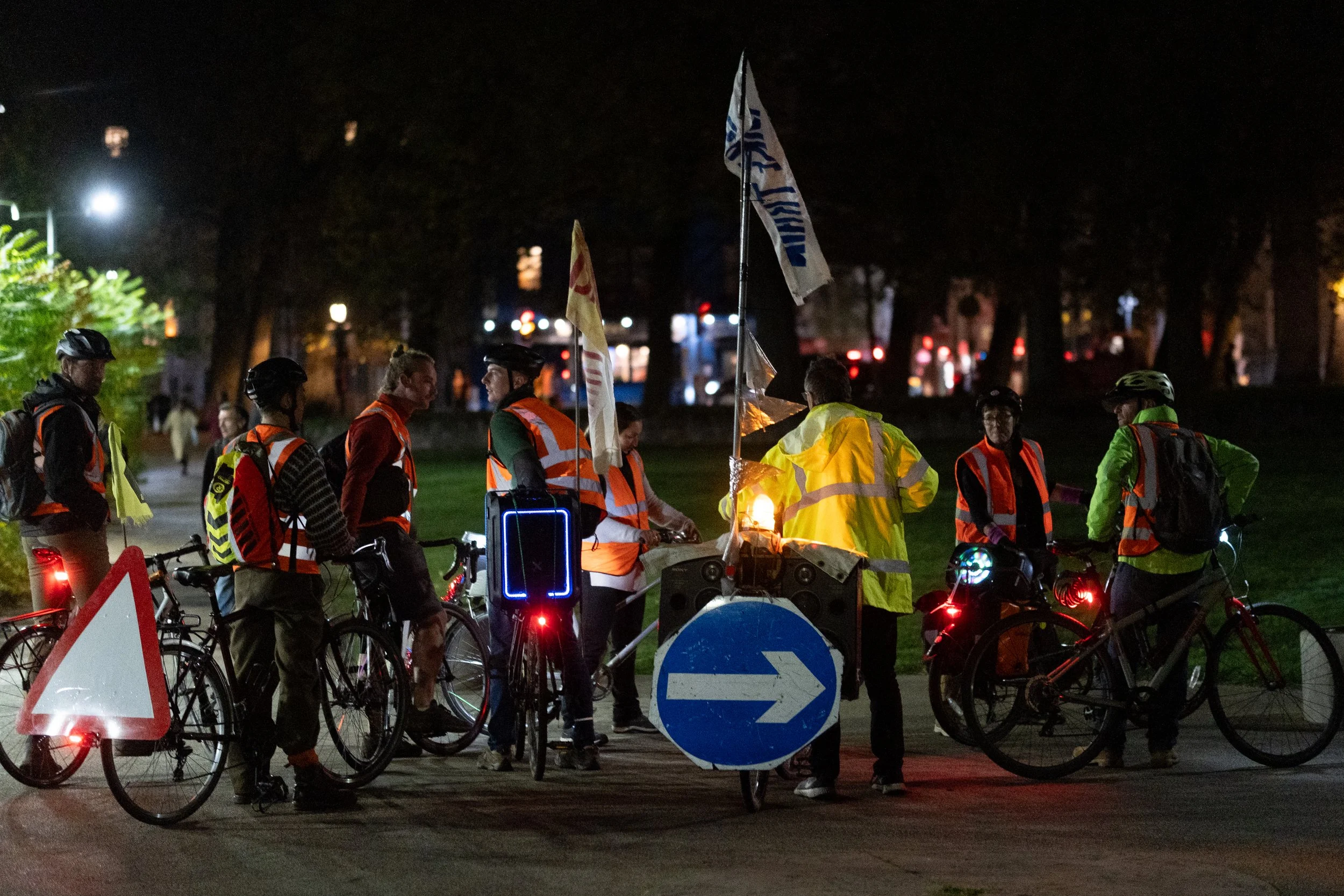 Group of cyclists wearing reflective gear gathered at night, with some holding flags, and a bicycle with a large blue arrow sign, campfire, and bicycle lights illuminating the scene.