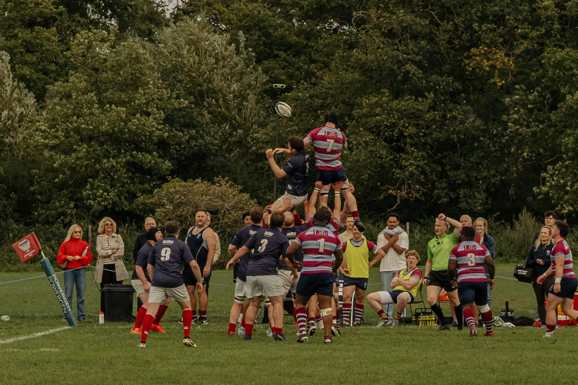 A rugby match in progress with players engaging in a line-out, lifting a player to catch the ball, as spectators watch on the sideline.