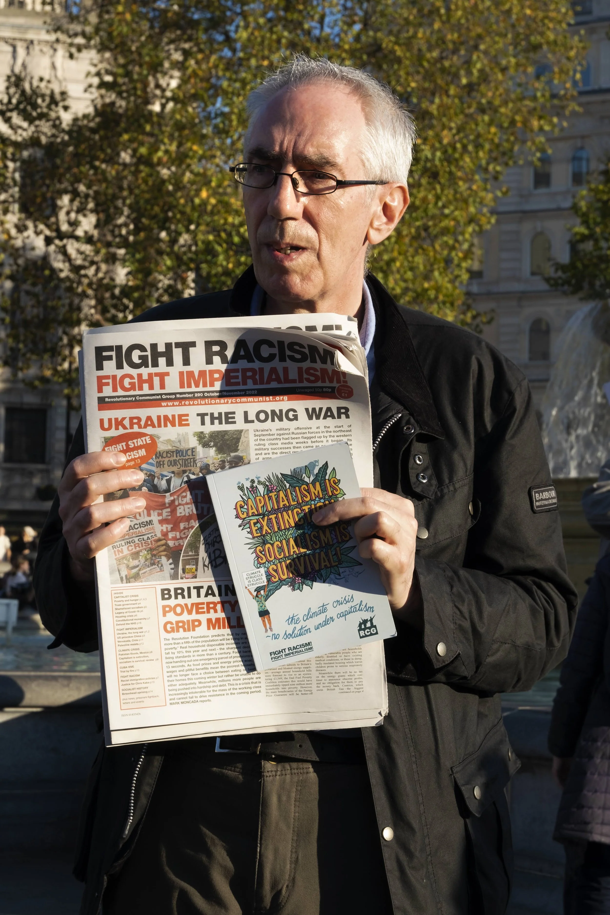 A man wearing glasses and a black jacket holding a newspaper and a flyer in an outdoor setting with trees in the background.