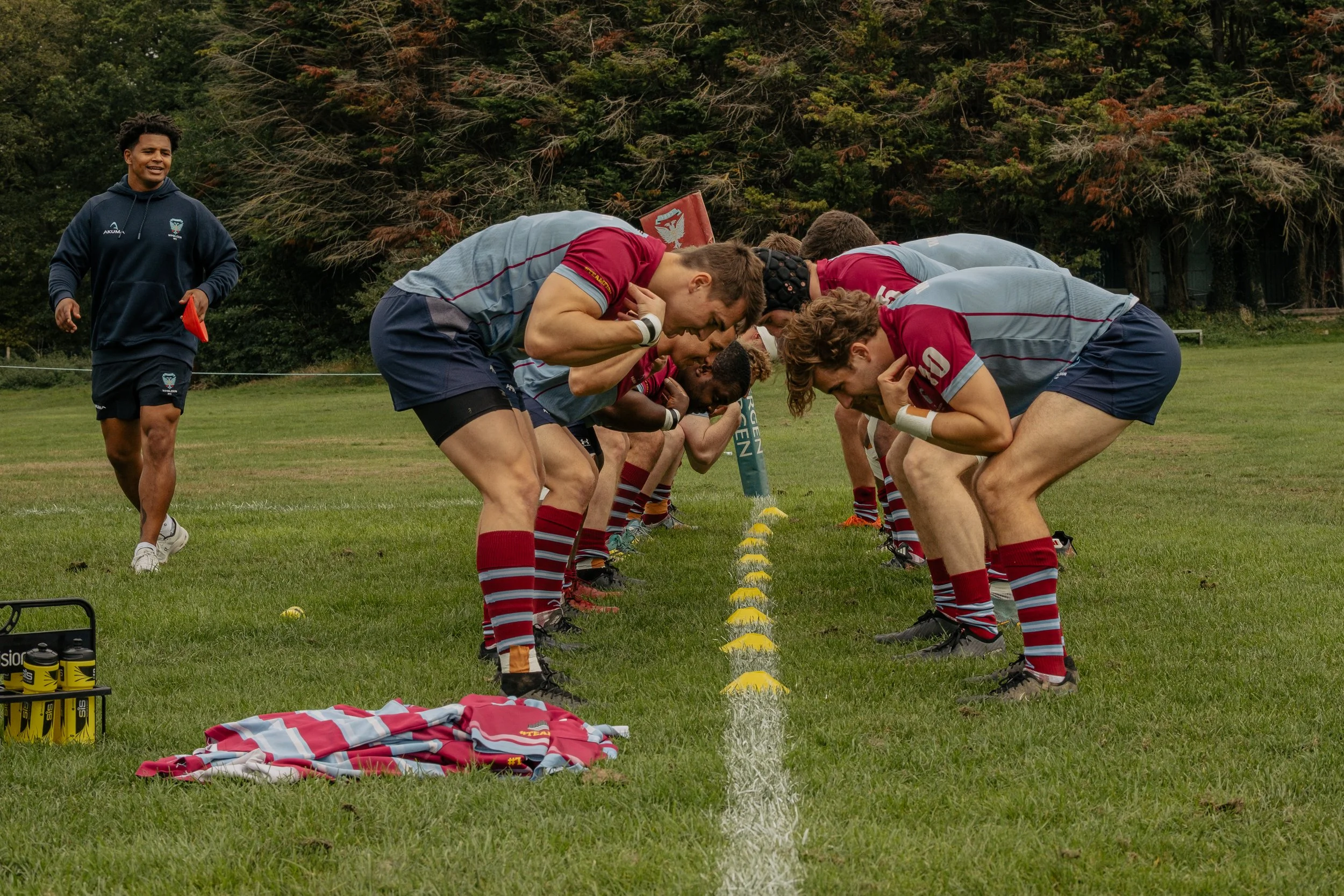 Rugby players practicing a scrum on a grassy field, with a coach observing in the background, and equipment like water bottles and jerseys nearby.