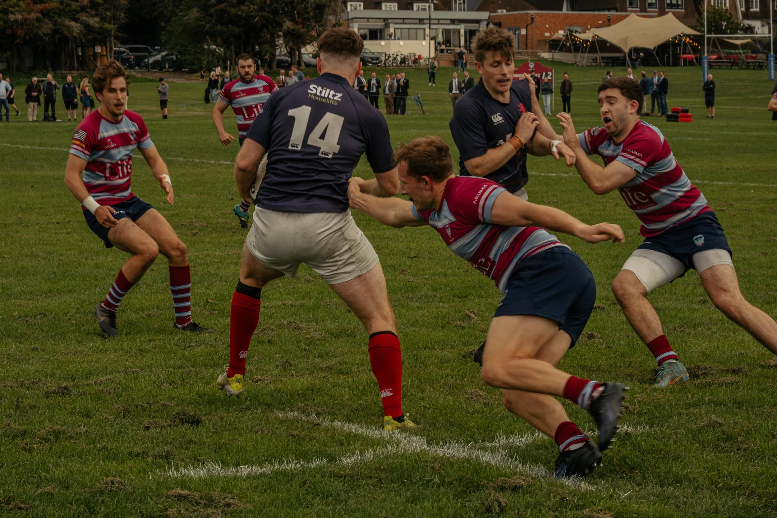 Rugby players competing for possession of the ball during a game on a grassy field with spectators and tents in the background.