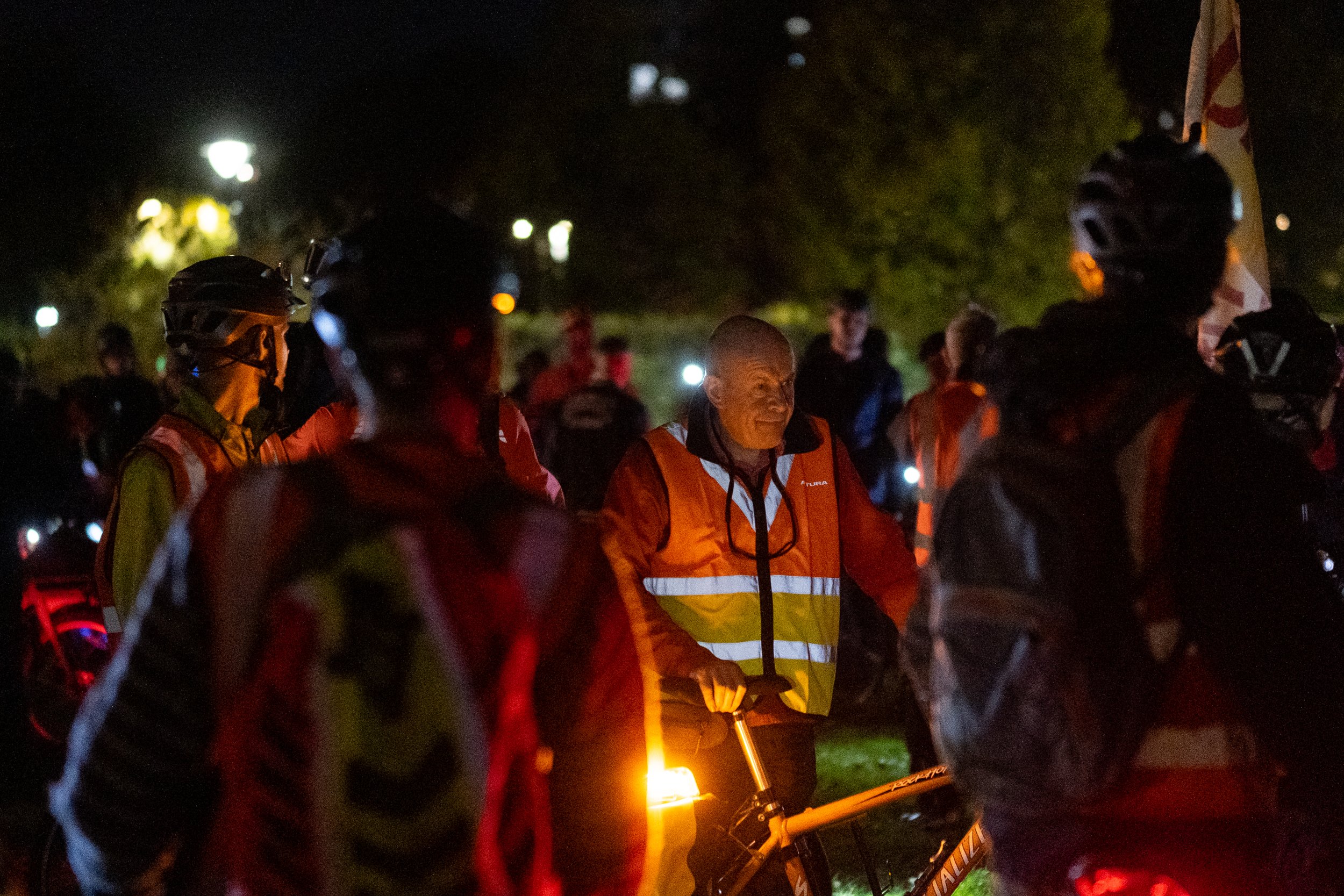 Group of people in high-visibility jackets and helmets gathered outdoors at night, with some holding bicycles and flashlights.