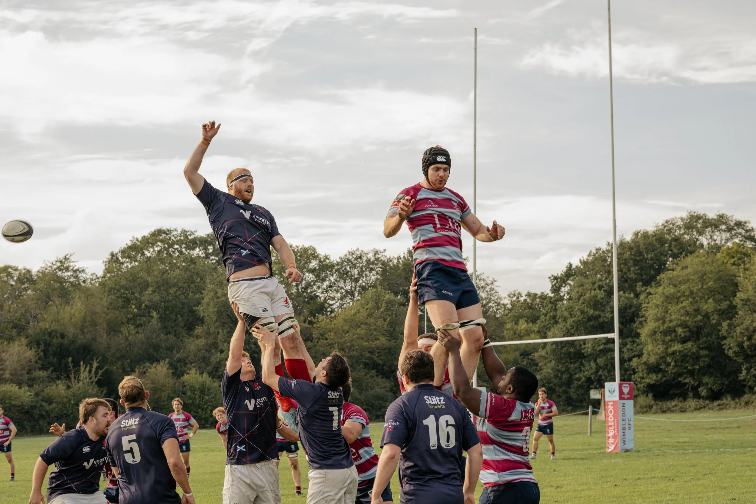 Rugby players competing in a lineout during a match, with players lifting a teammate to catch the ball, on a grassy field with trees in the background.