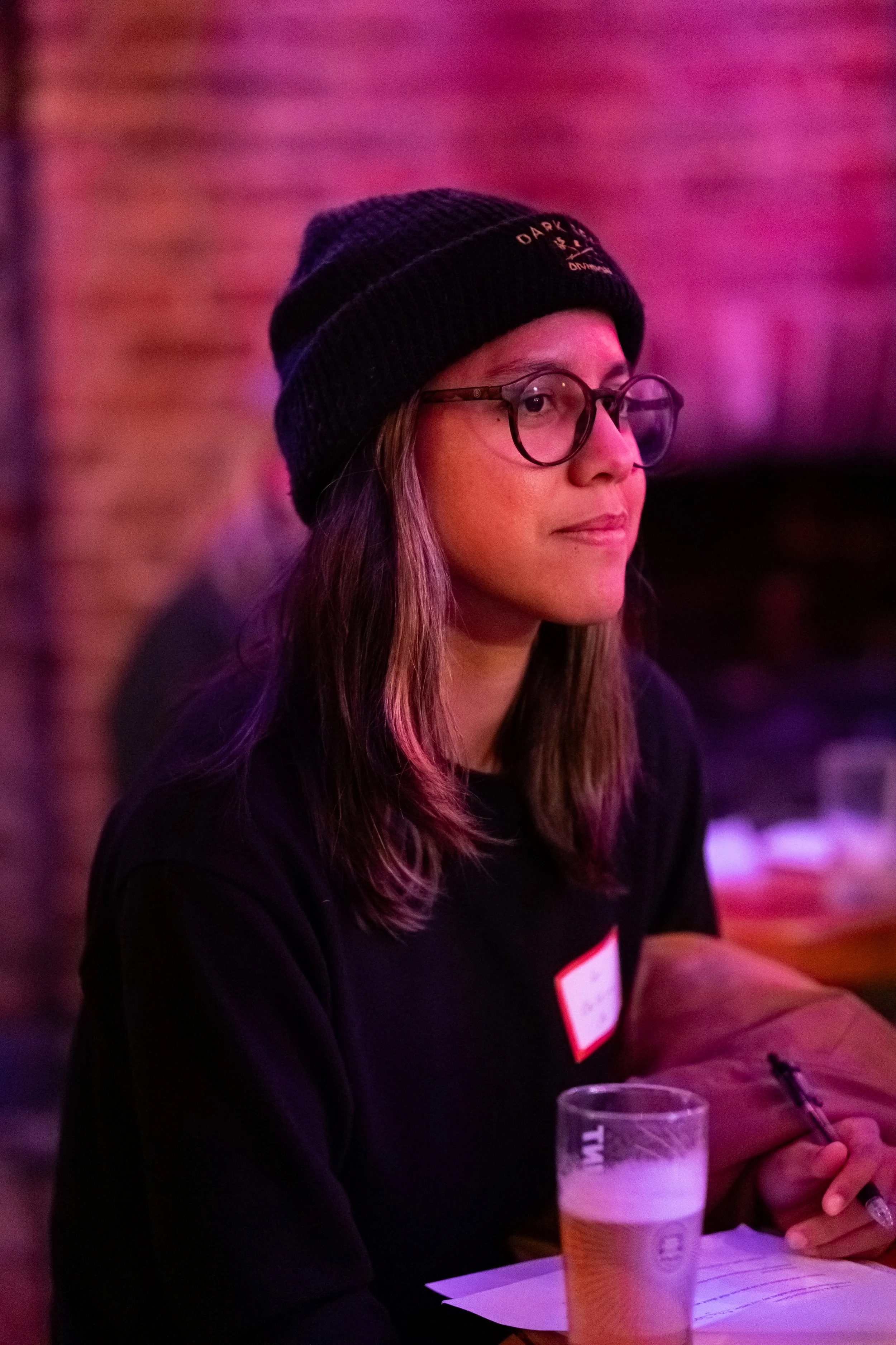 A young woman with glasses and long hair, wearing a black beanie, sitting at a table with a glass of beer and a notepad, in a dimly lit setting with pink and purple lighting.
