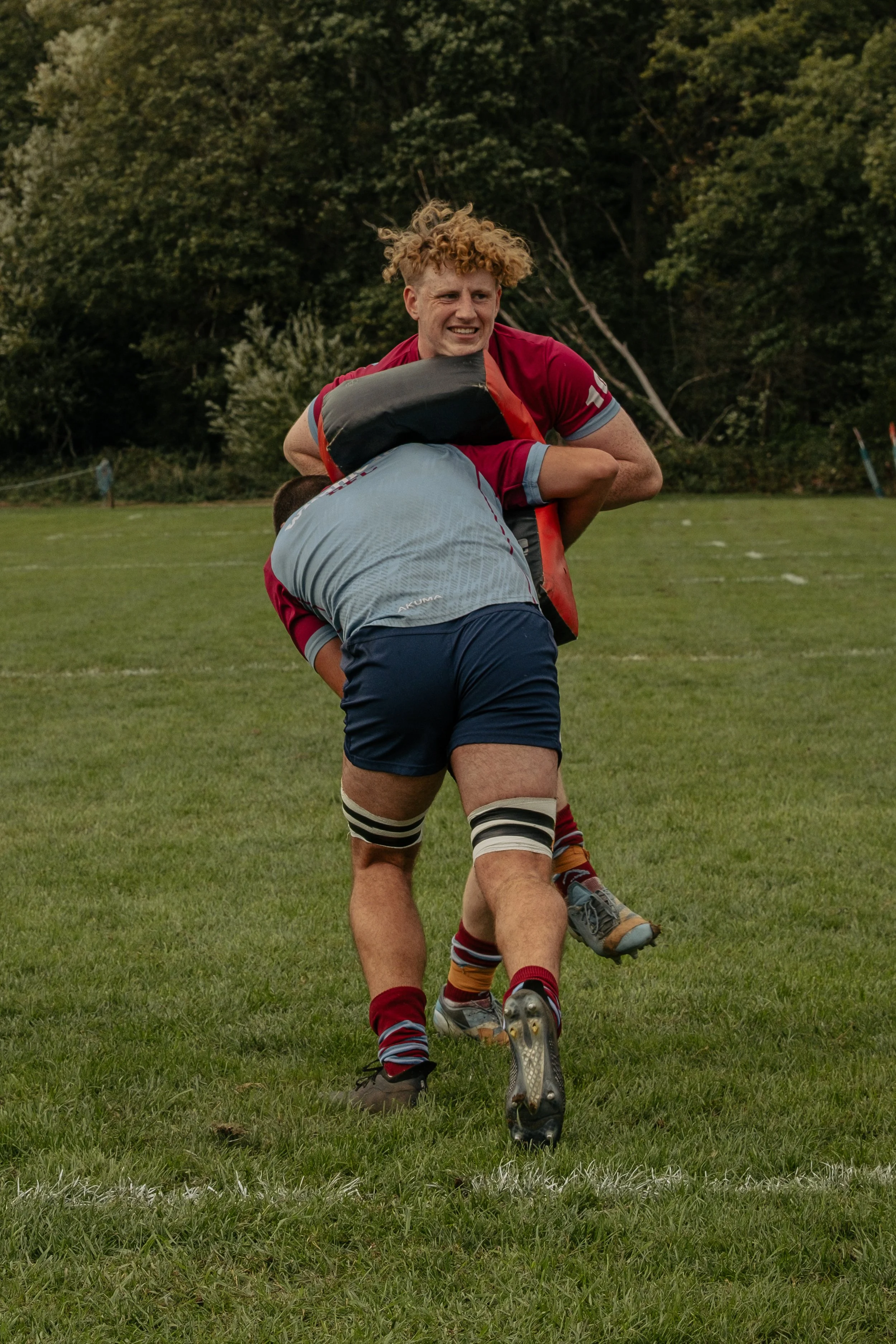 Two rugby players in a tackle during a game on a grass field with trees in the background.