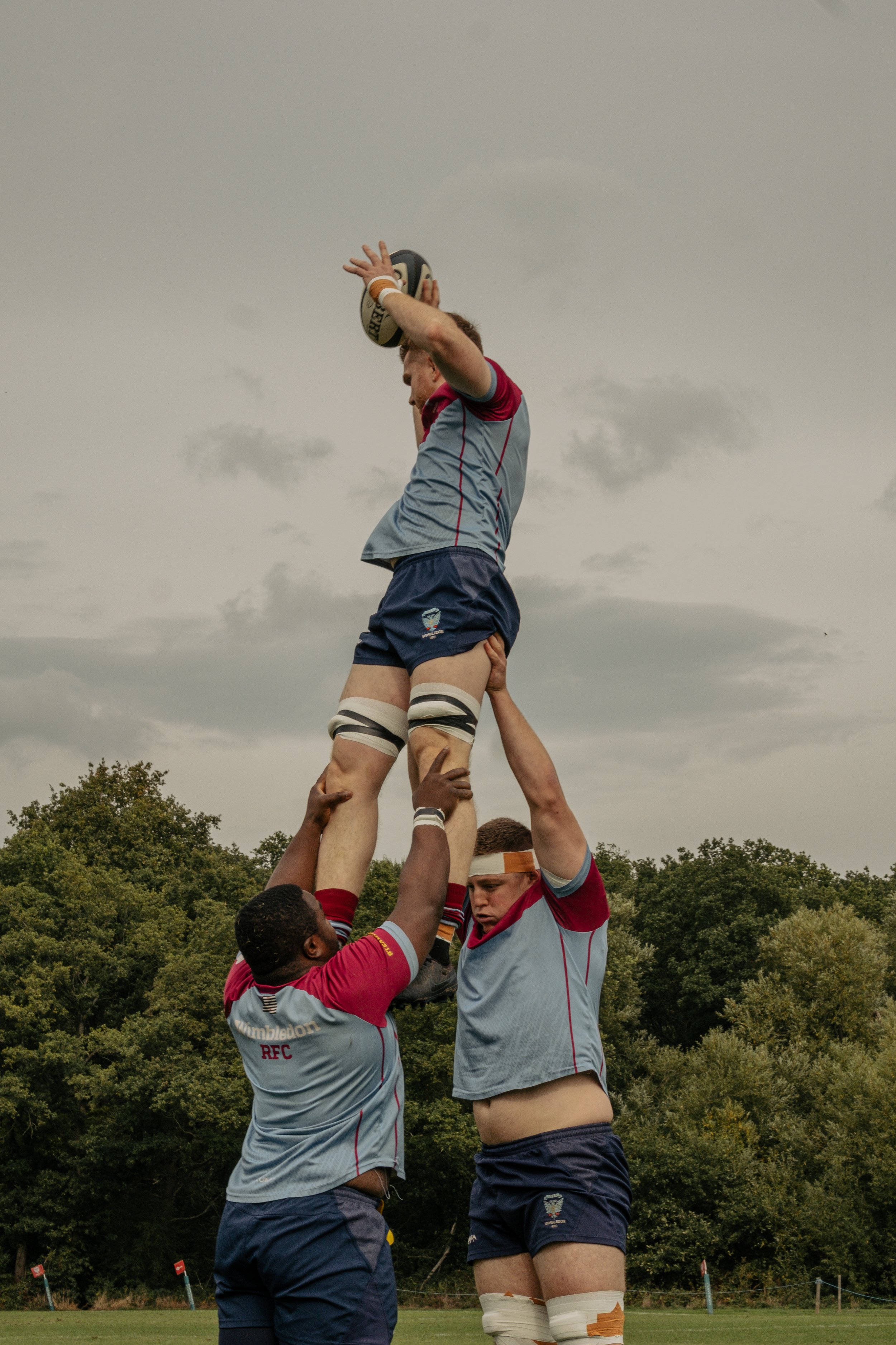 Rugby players performing a lineout lift during a match, with one player holding the rugby ball above his head, outdoors on a cloudy day.