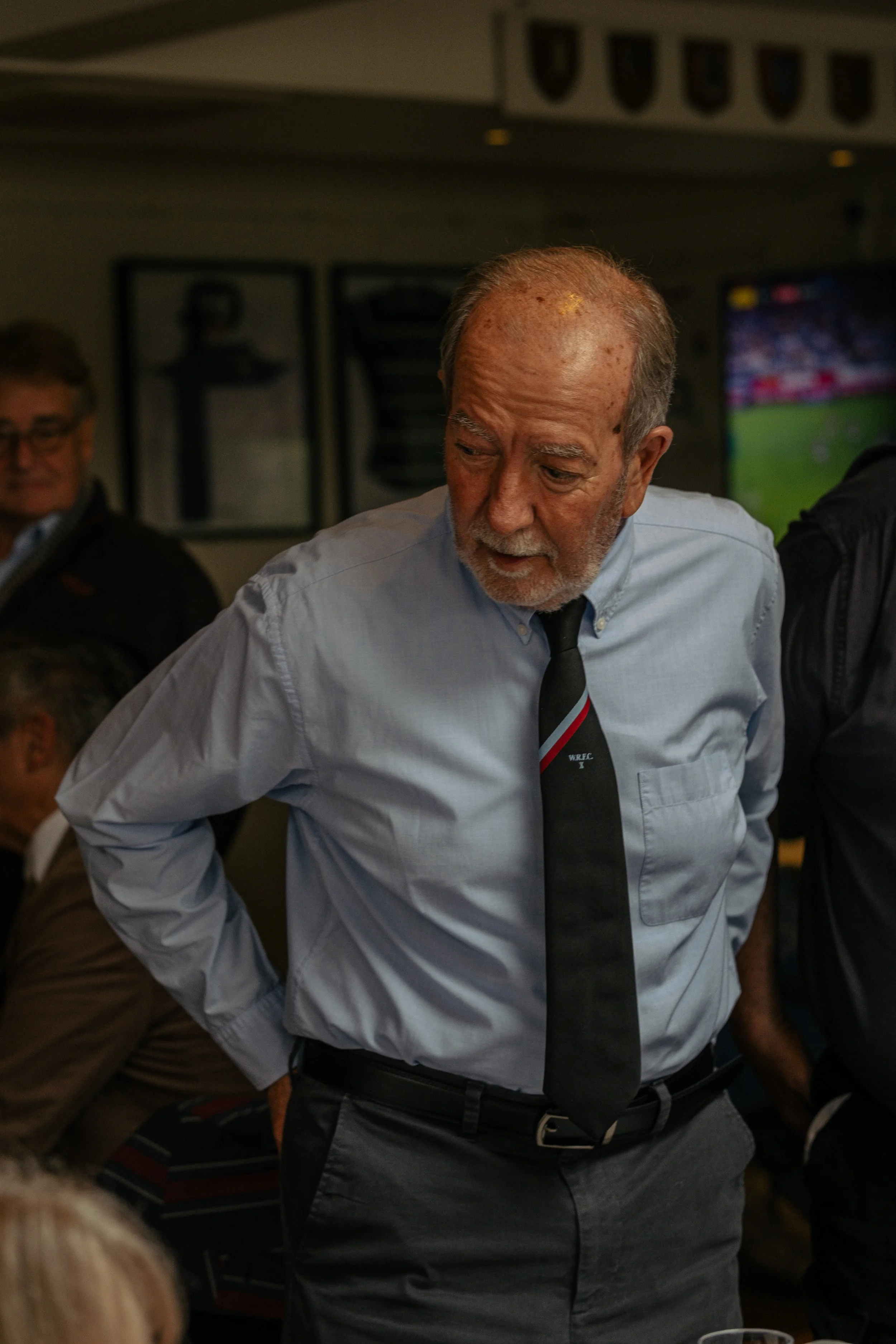 An older man with a gray beard and hair, wearing a light blue button-up shirt, a black tie, and dark pants, standing with his hands on his hips in a room with framed pictures and a television in the background.