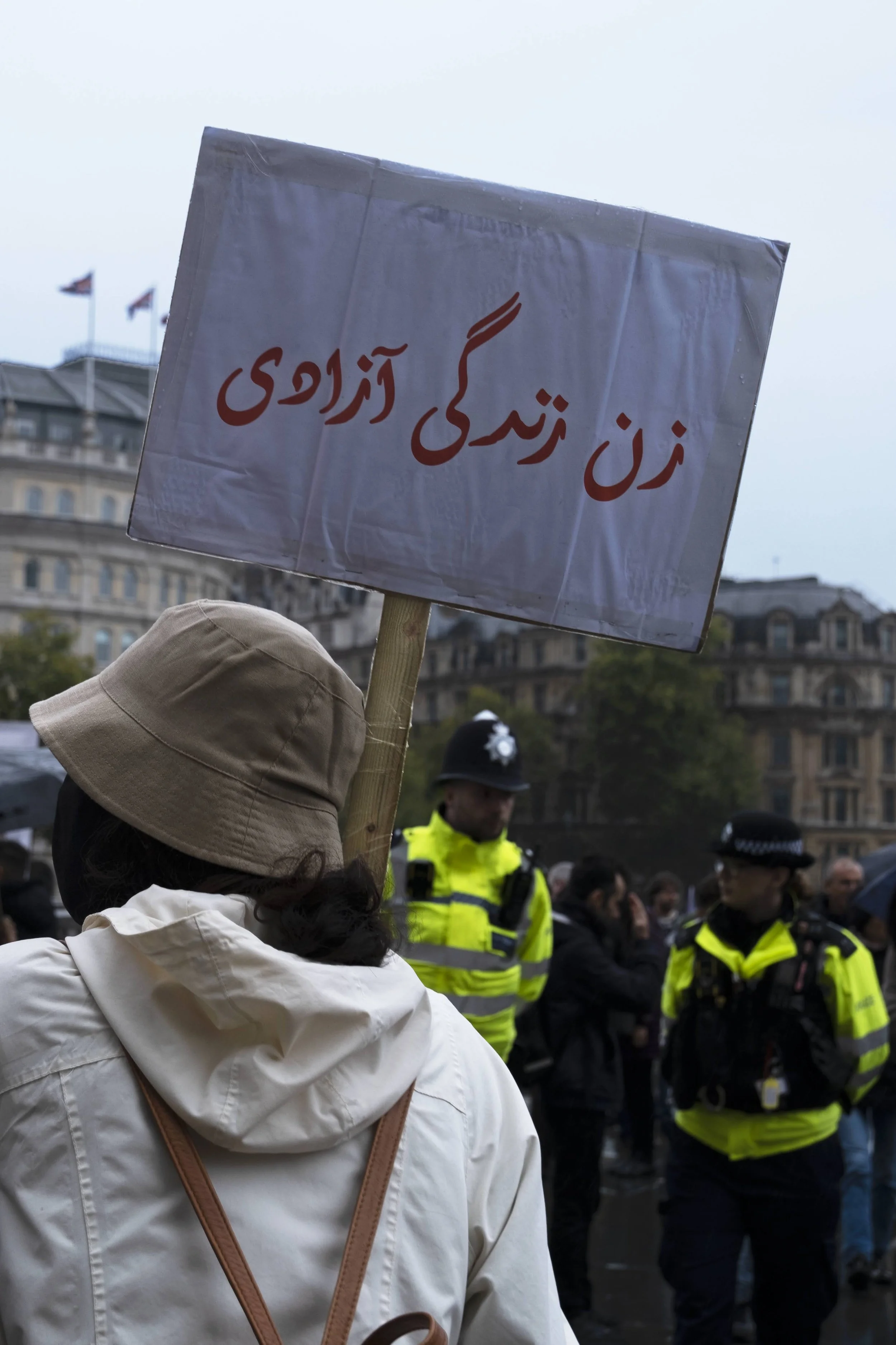 Person holding a sign with Persian writing during a protest, police officers and a crowd in the background in an urban setting.