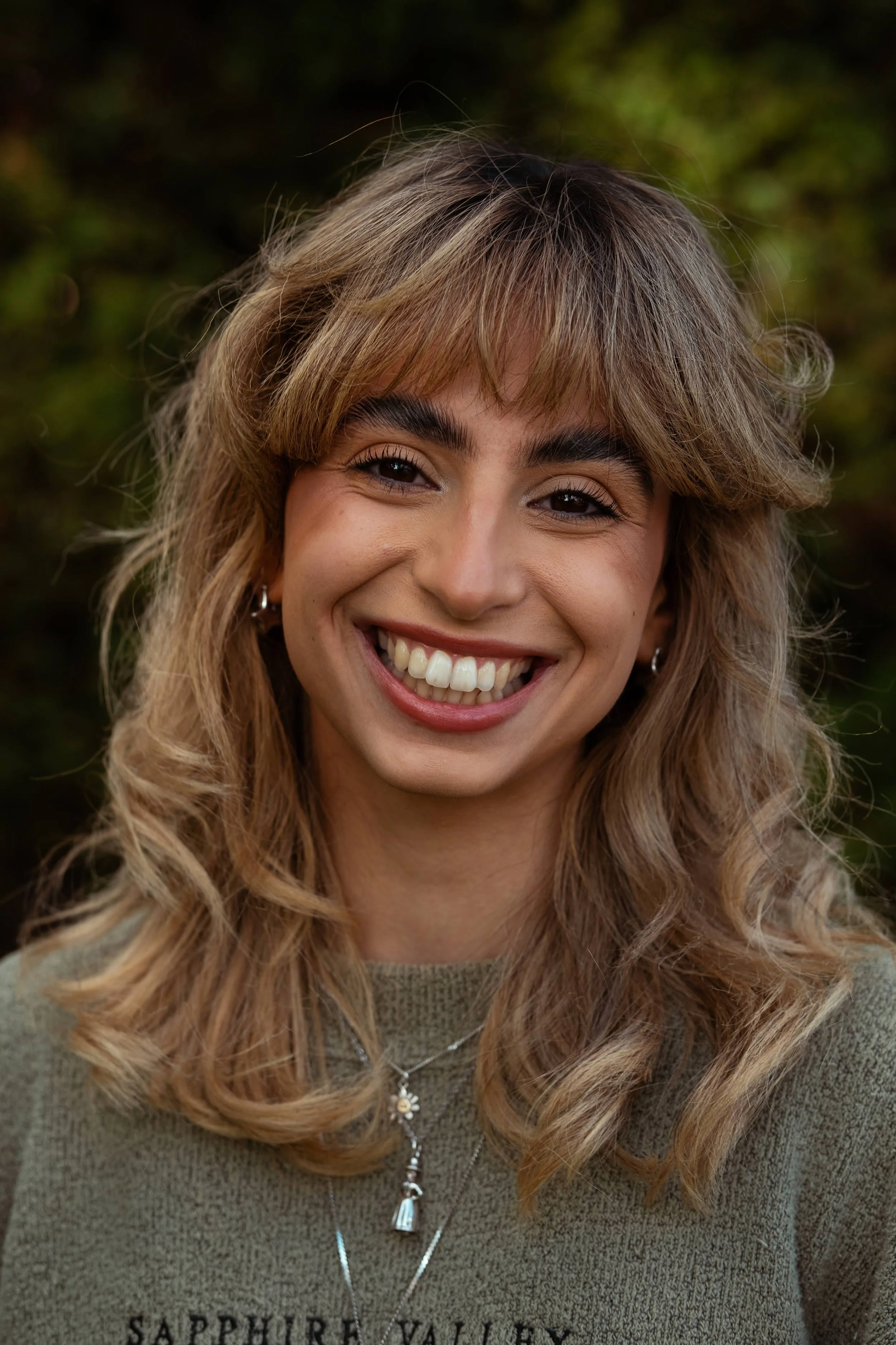Close-up of a smiling woman with wavy blonde hair outdoors, background of foliage, wearing a beige top with 'SAPPHIRE VALLEY' text, and layered necklaces.