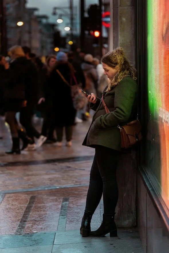 A woman standing on a city sidewalk at night, leaning against a building wall and looking at her phone. She is dressed in a dark green coat, black pants, and black heeled boots, with a brown purse slung over her shoulder. There are blurred pedestrian