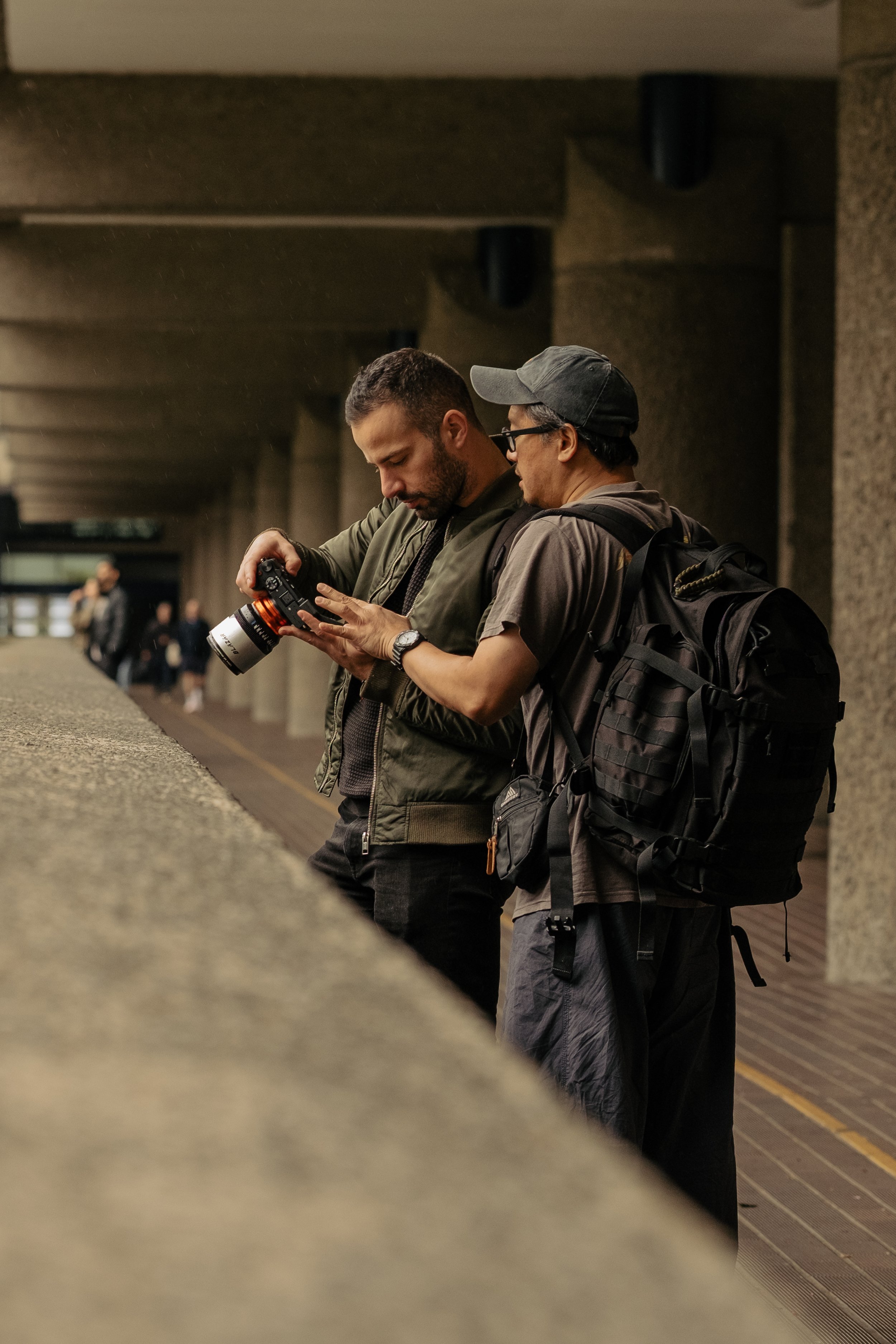 Two men looking at a camera on a ledge in a tunnel or underpass, one with a backpack, with other people in the background.