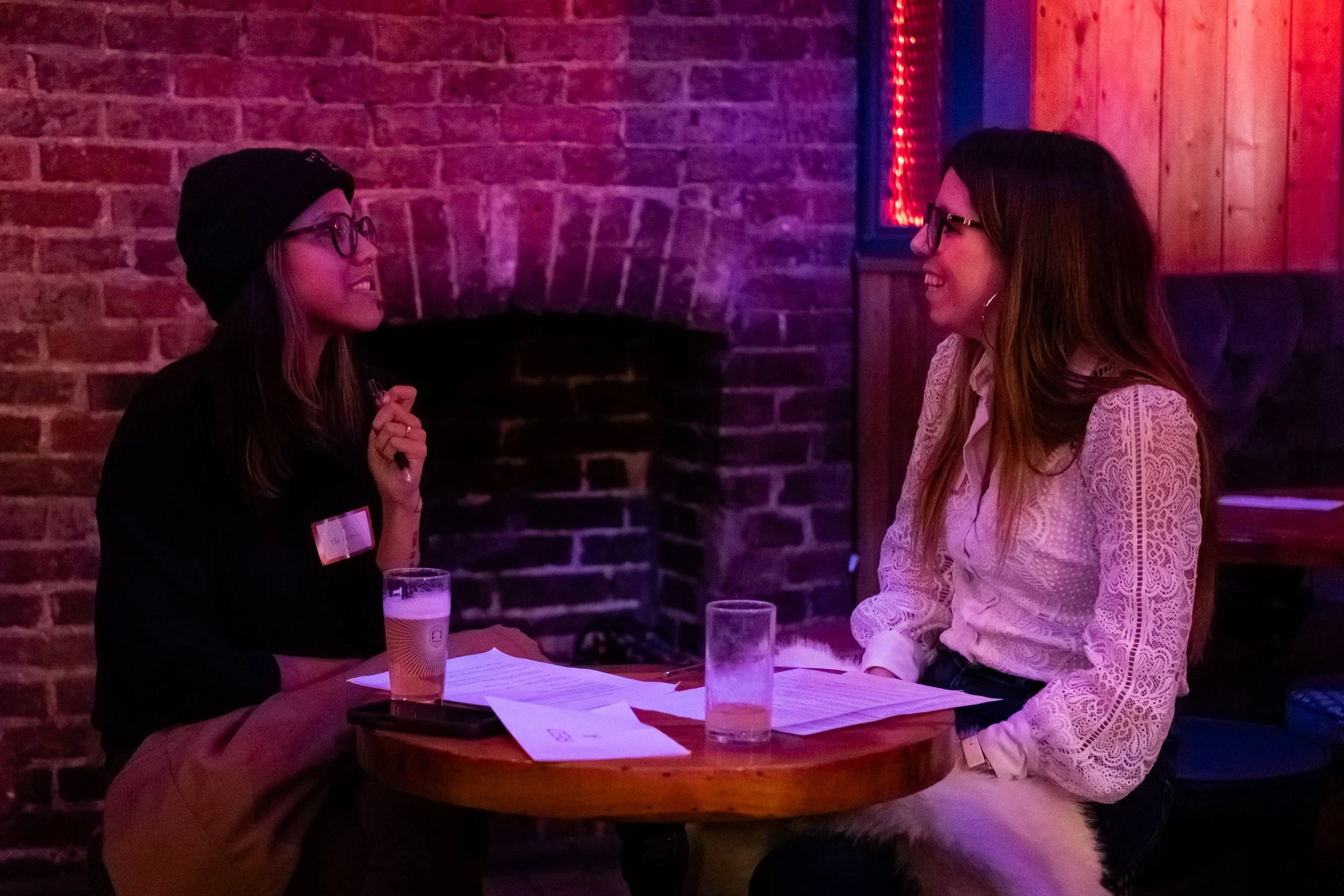 Two women sitting at a round wooden table in a dimly lit bar, engaged in conversation. The woman on the left is wearing a black beanie, glasses, black shirt, and tan pants with a glass of beer in front of her. The woman on the right is wearing a whit