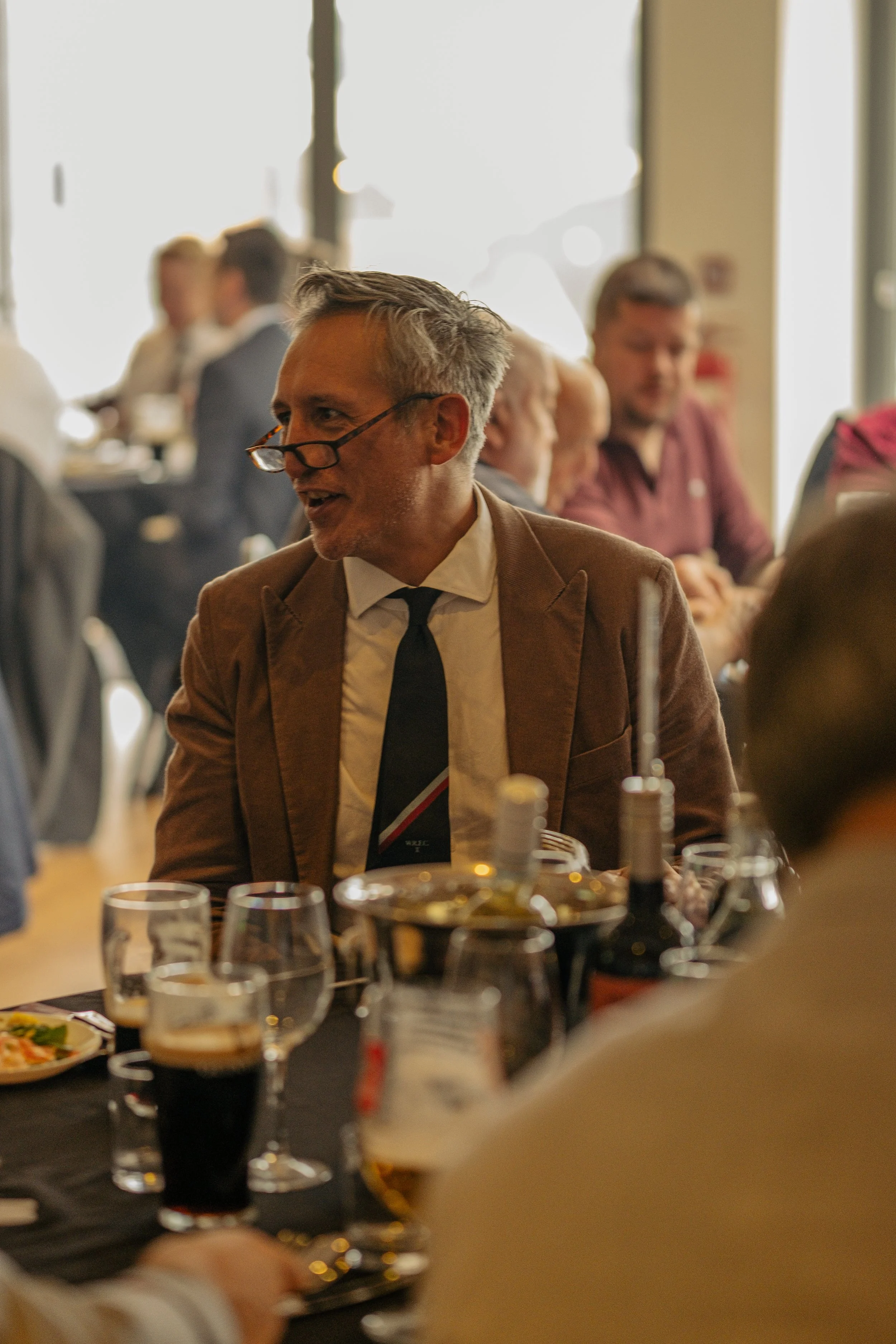 A middle-aged man with glasses, dressed in a brown blazer, white shirt, and black tie, is talking at a formal dinner event with multiple glasses of beverages and bottles on the table. Others are visible in the background.