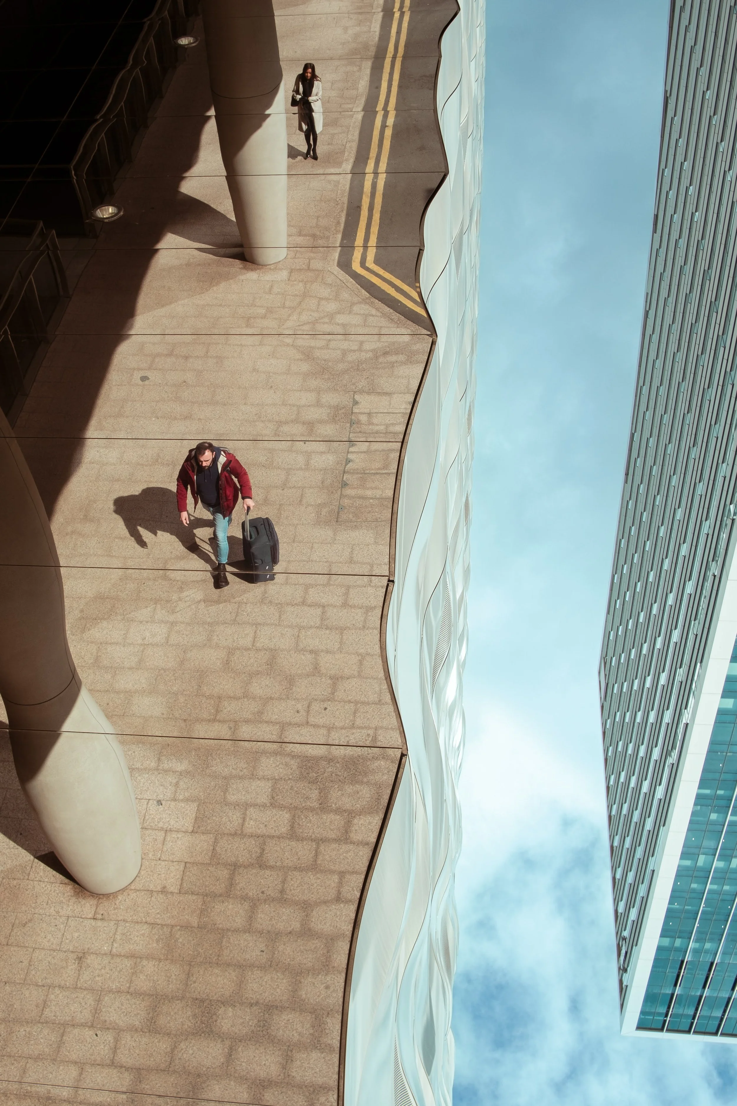 Reflection of two people walking on a sidewalk next to modern buildings, with the sky showing in the reflection.