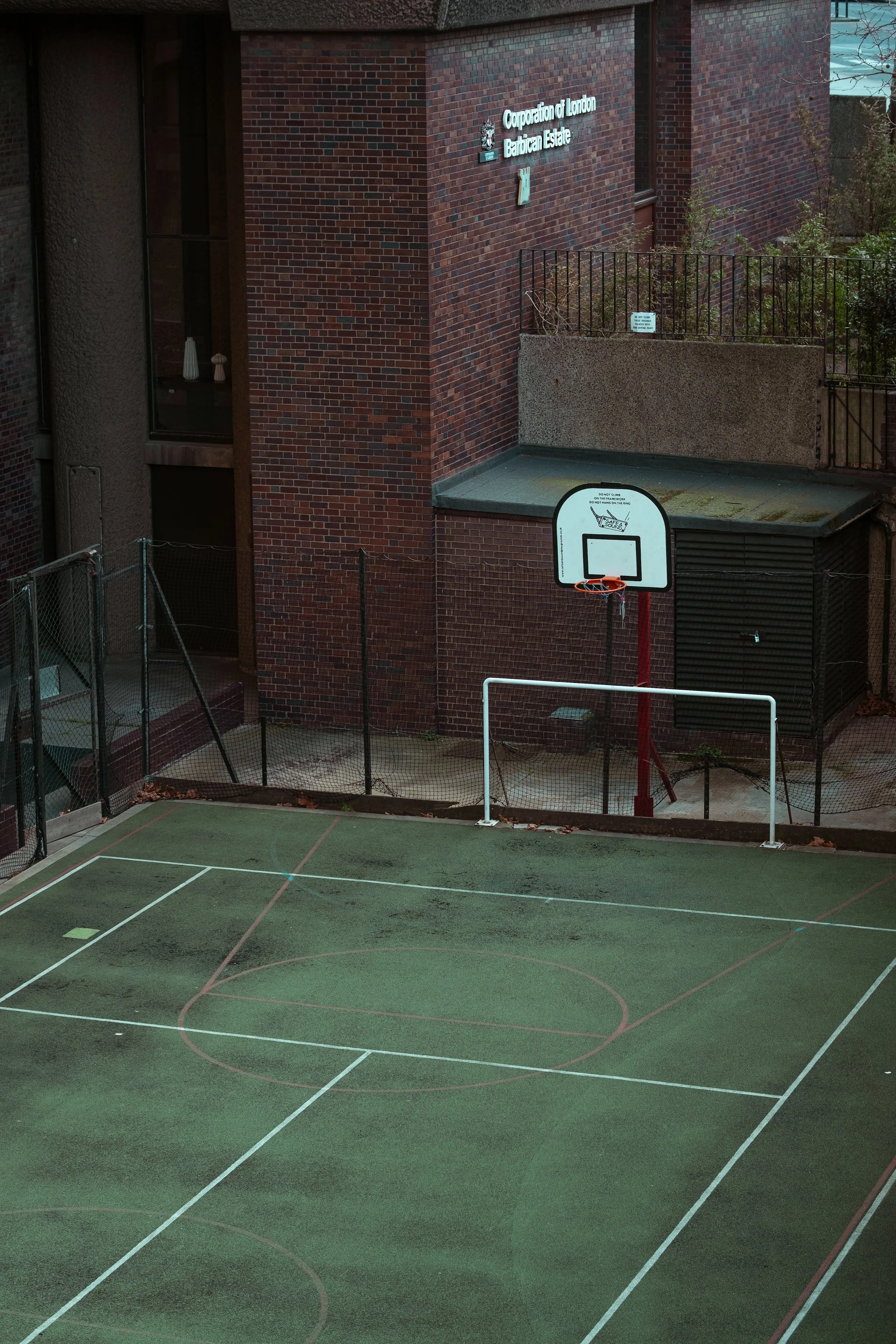 Empty outdoor basketball court with a hoop, small goal, and surrounding fencing, adjacent to a brick building with a sign that reads 'Corporation of London Barbican Estate'.
