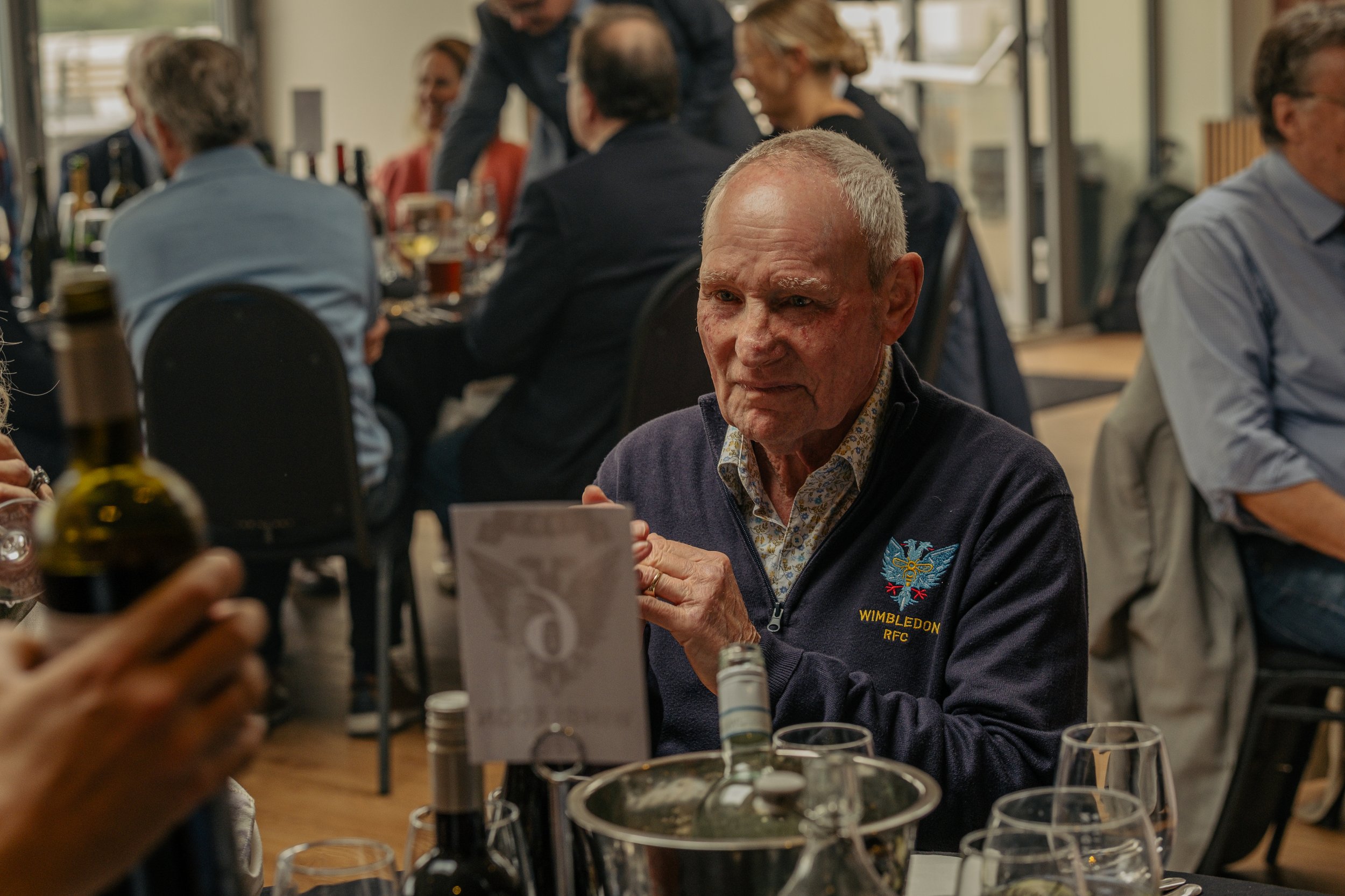 An elderly man wearing a Wimbledon RFC jacket sitting at a table during a social event, with wine glasses and bottles on the table, and other people in the background.