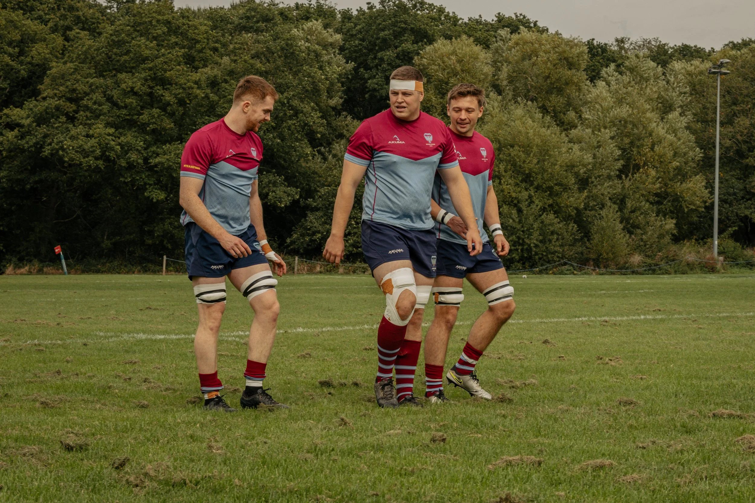 Three rugby players with supportive expressions, wearing matching maroon and light blue jerseys, navy shorts, and striped socks, walking on a grassy field with trees in the background.