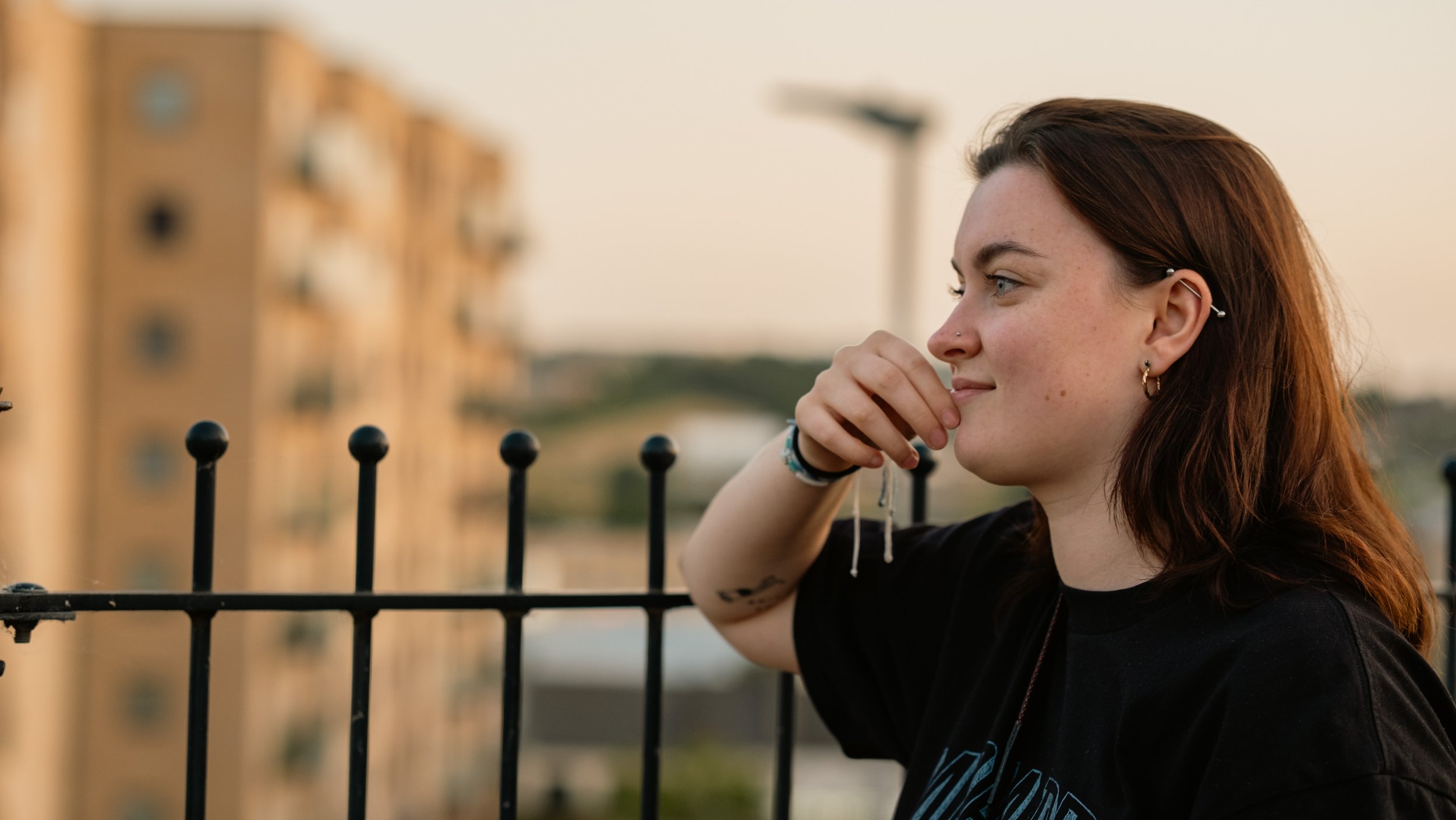 A young woman with reddish-brown hair and multiple earrings, smiling thoughtfully while leaning on a black metal fence, with a blurry building and sunset in the background.