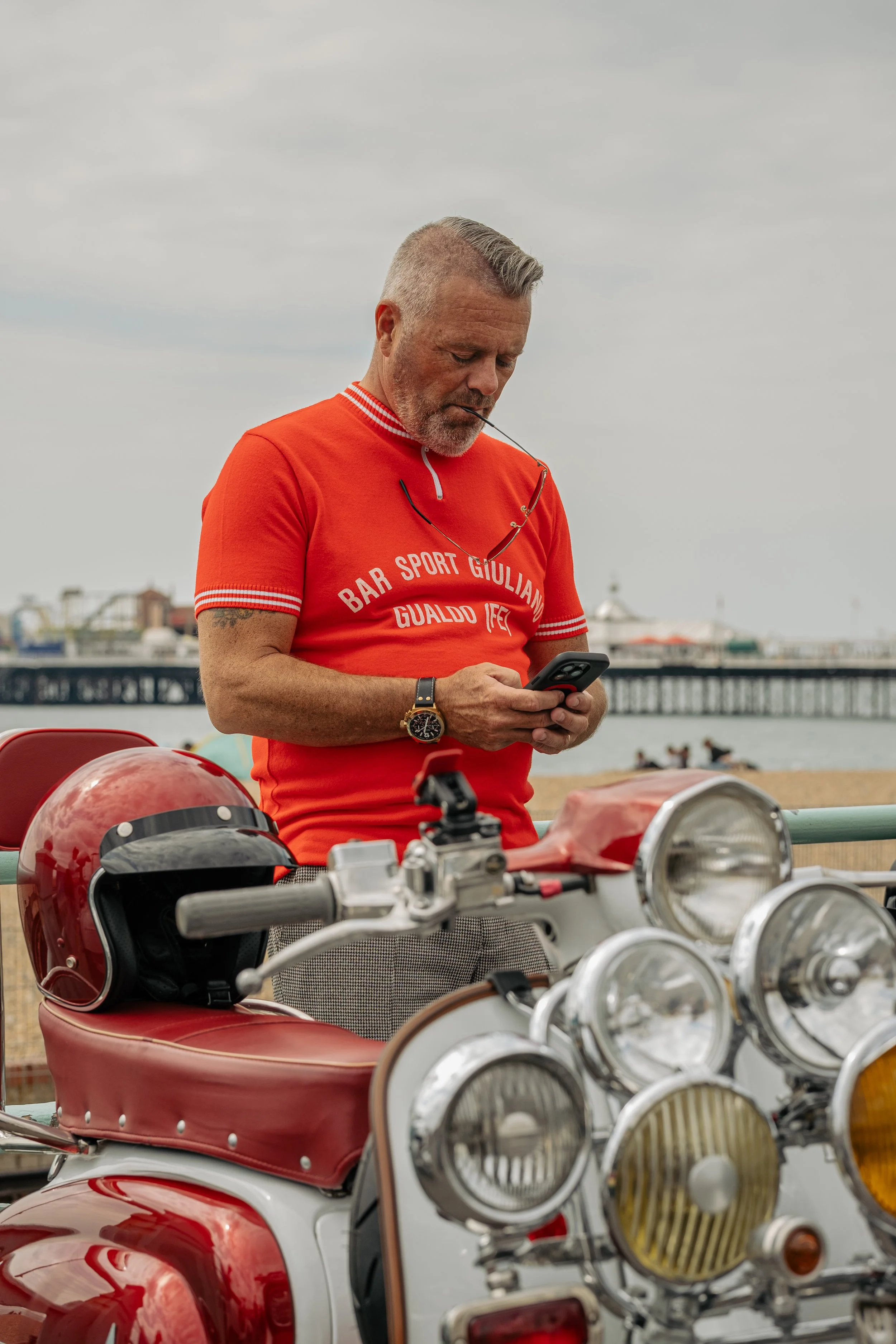 A middle-aged man with grey hair and facial hair wearing a red T-shirt, standing next to a vintage red motorcycle on a beach. He is looking at his phone with sunglasses hanging from his shirt collar and a wristwatch on his left wrist. A red helmet is