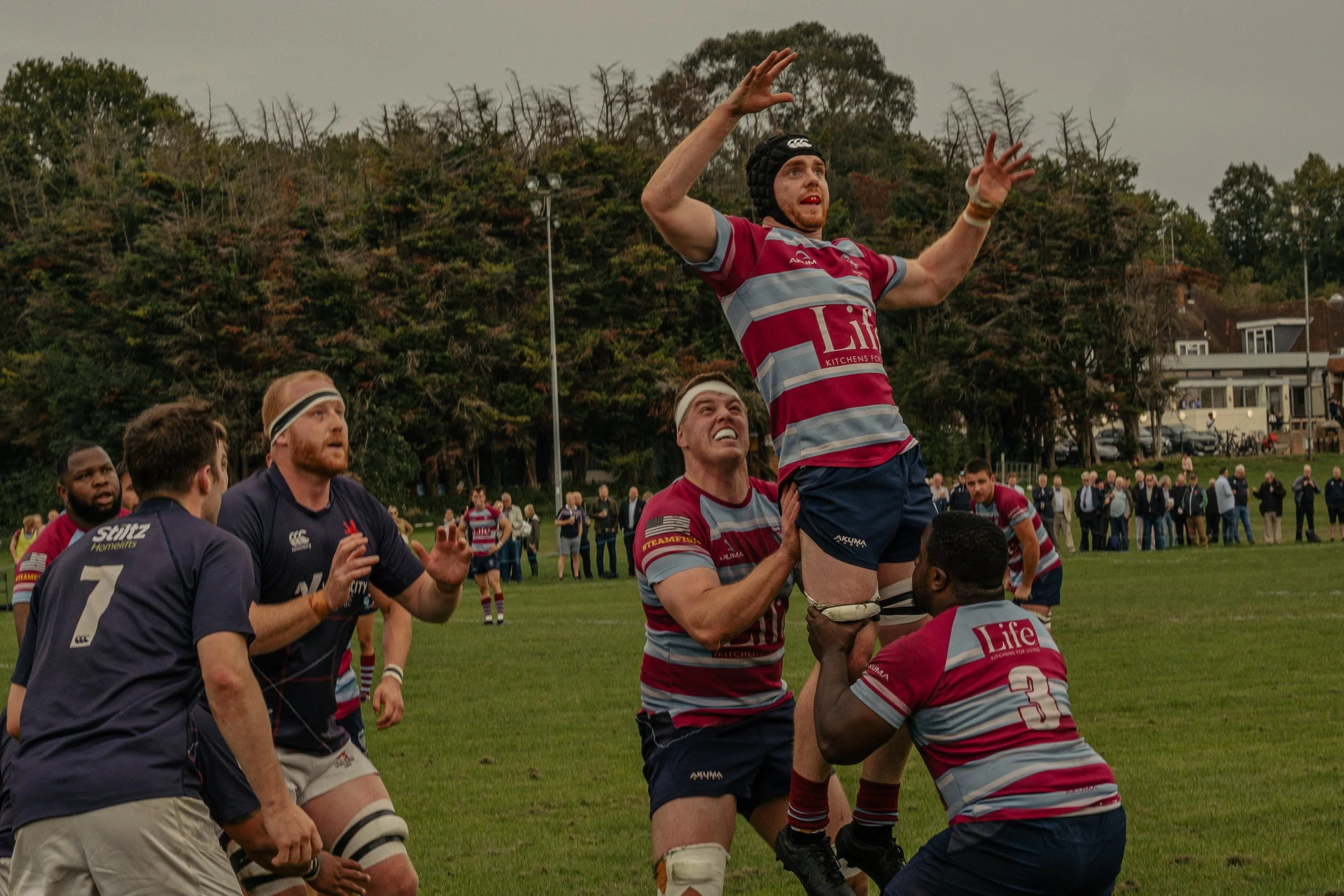 Rugby players in maroon and blue uniforms form a lineout to catch the ball during a match on a grassy field, with spectators in the background and trees on a cloudy day.
