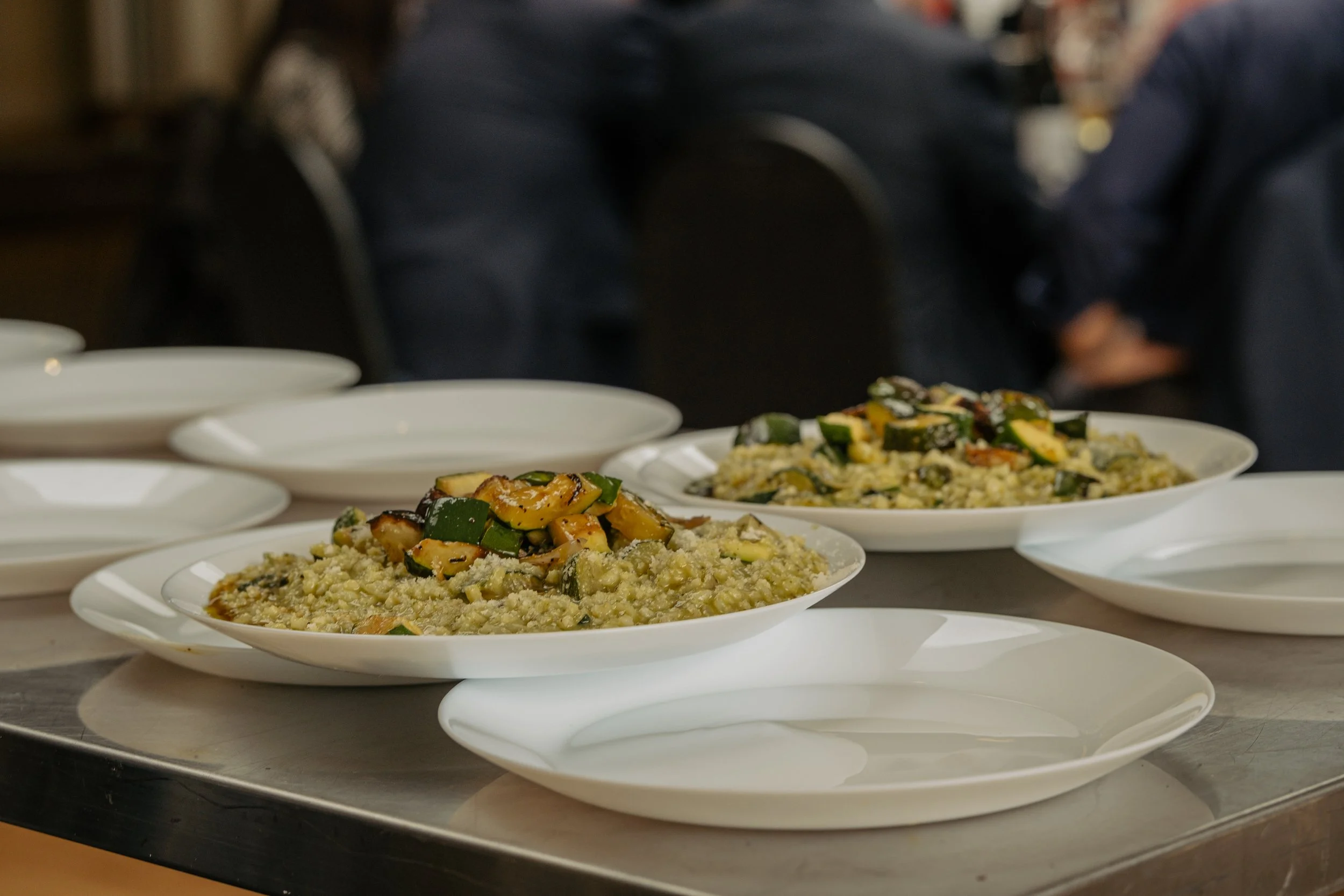 Two plates of cooked zucchini and what looks like quinoa or couscous, in a restaurant setting with people sitting at a table in the background.