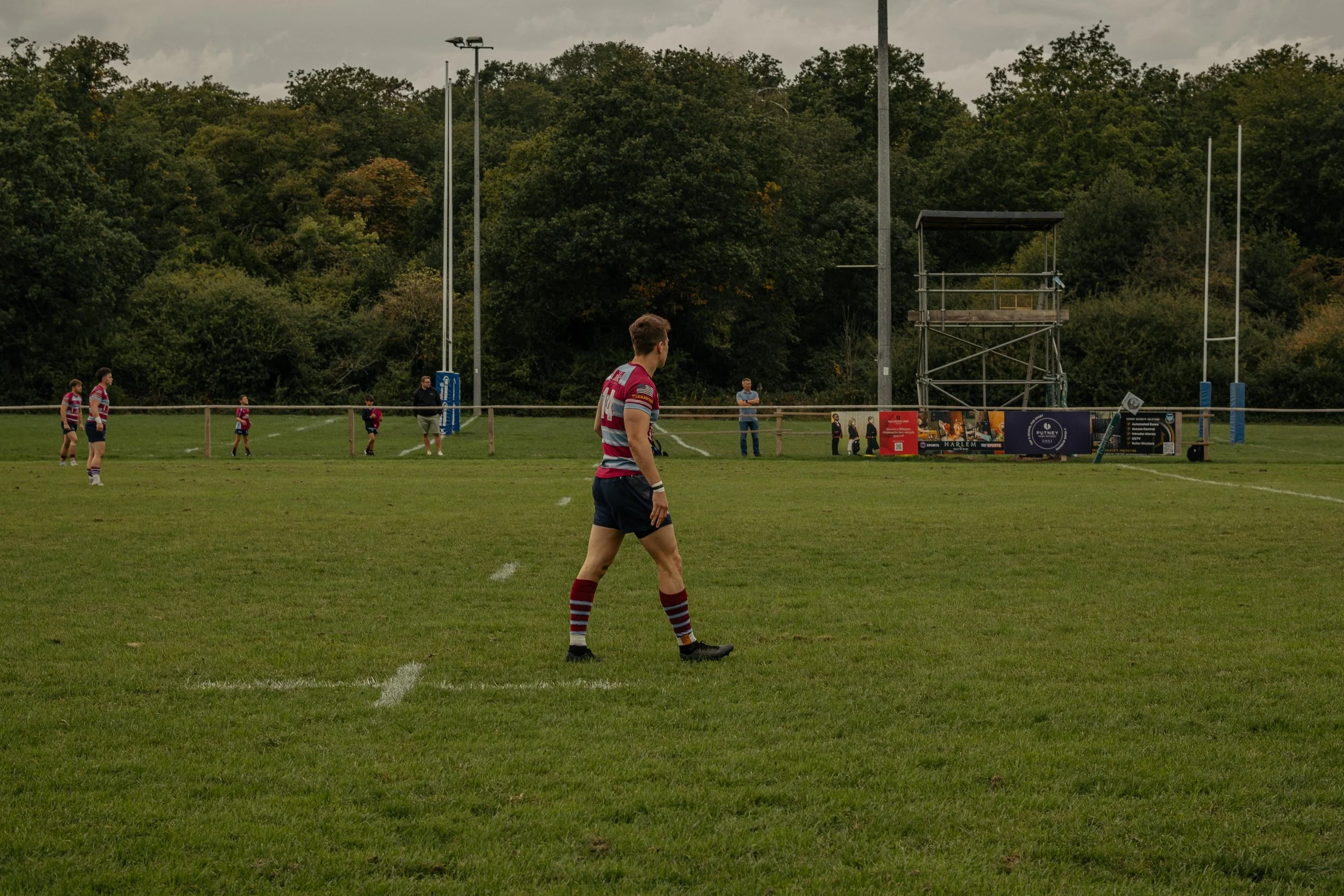 Rugby players warming up on a grassy field during daytime with a tree-covered hill in the background, rugby goalposts, and a small group of spectators.