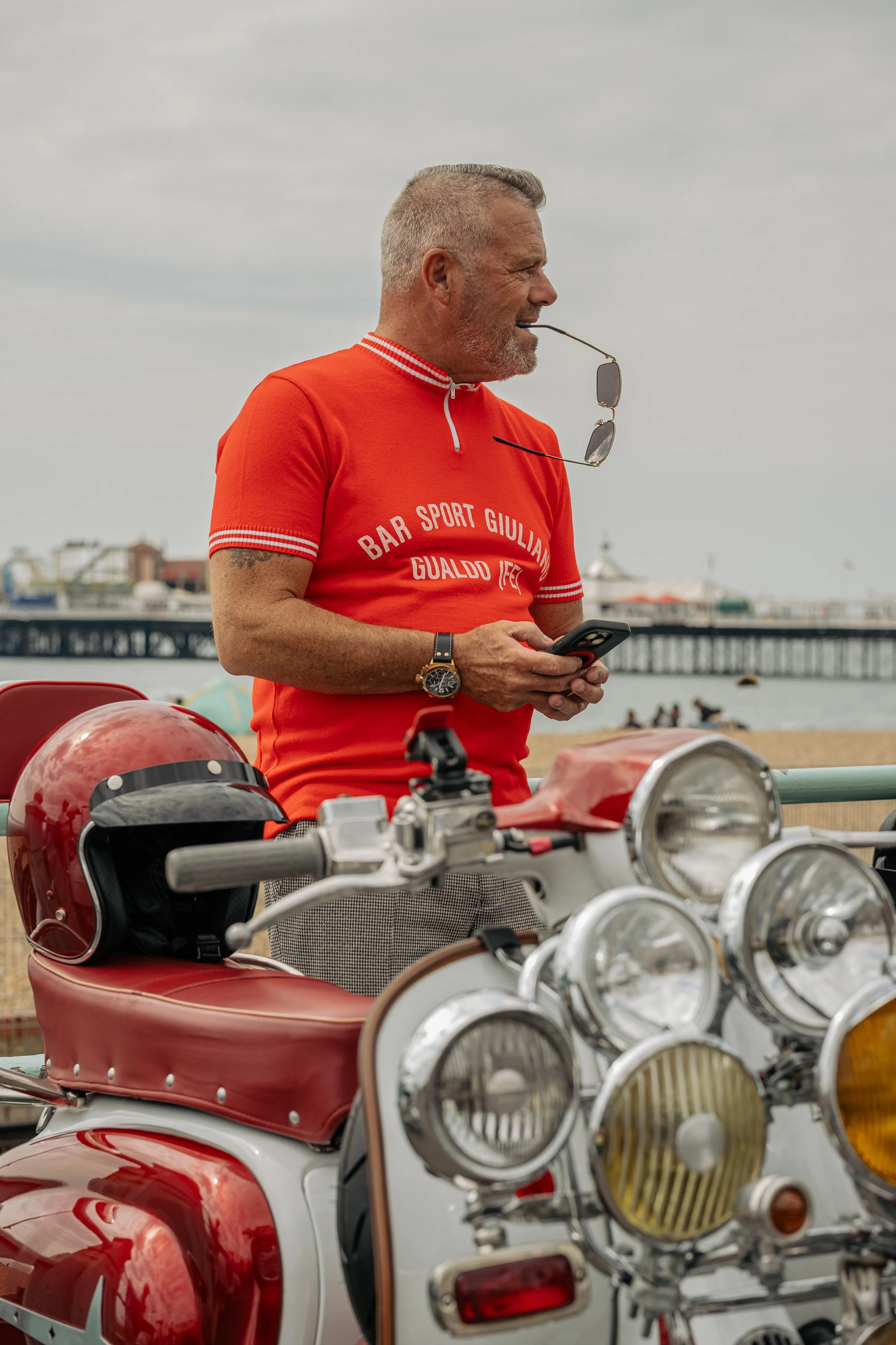 A man in a red shirt with the words "Bar Sport G Giuliani Gualdo IEG" is standing near a vintage motorcycle at the beach, holding a smartphone with sunglasses hanging from his shirt, with a bridge and people in the background.