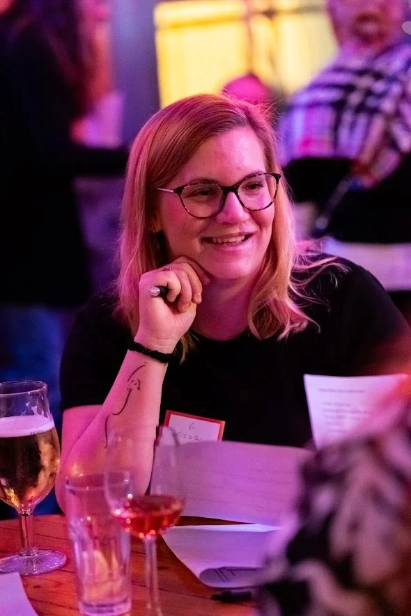 A young woman with glasses and red hair, smiling while sitting at a table with drinks, in a lively, colorful setting with other people in the background.