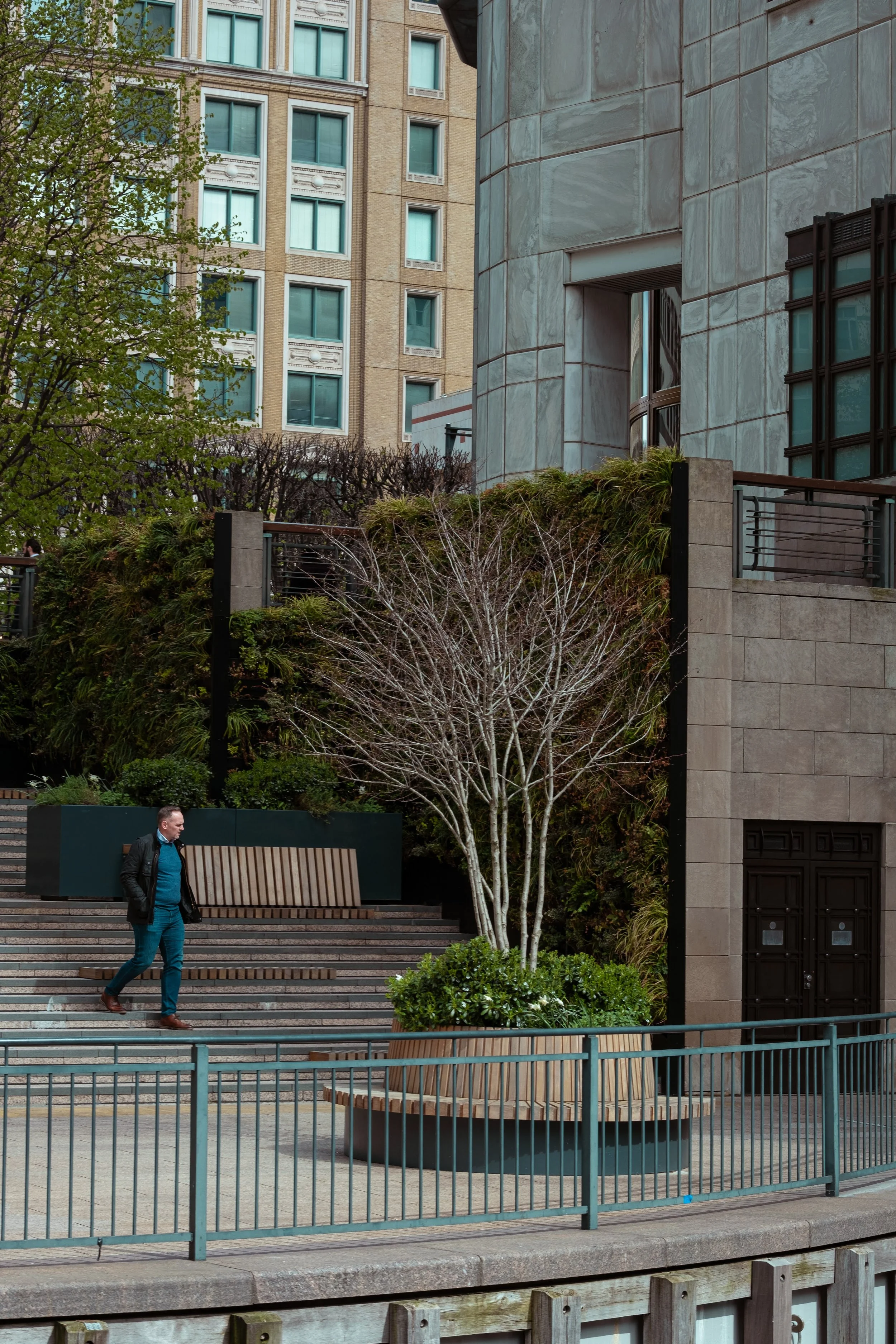 A man walking down stairs outside a modern building with trees and lush greenery, metal railing in foreground, and multi-story urban buildings behind.