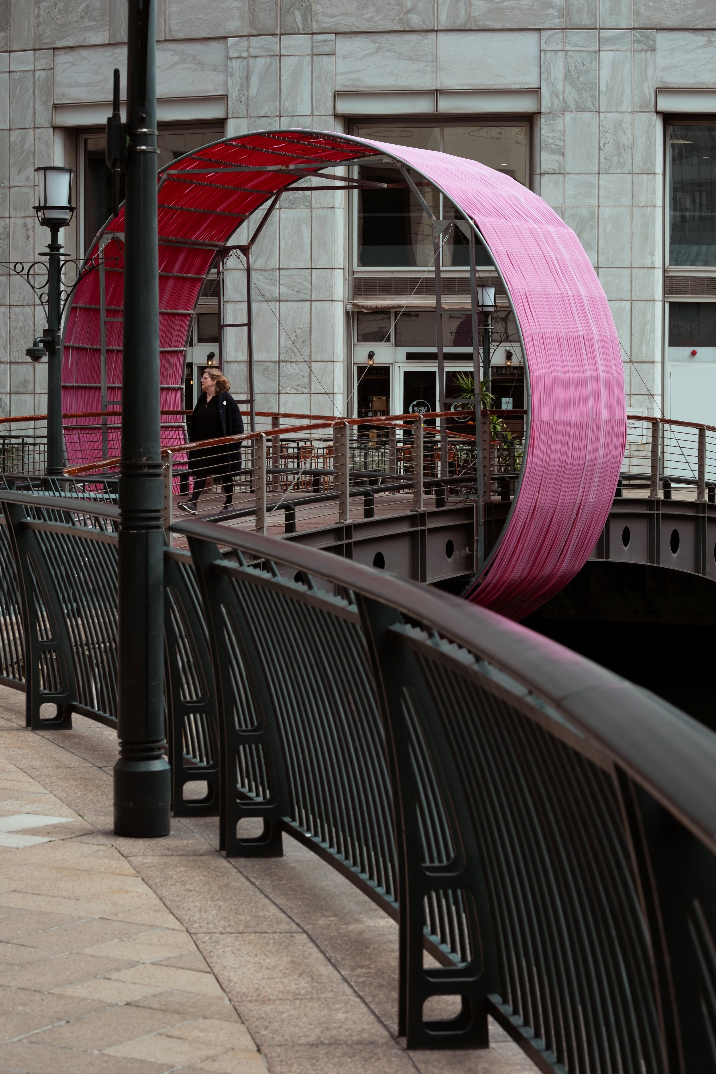 A person walking on a bridge with a pink, curved, semi-circular architectural structure behind them in an urban setting.