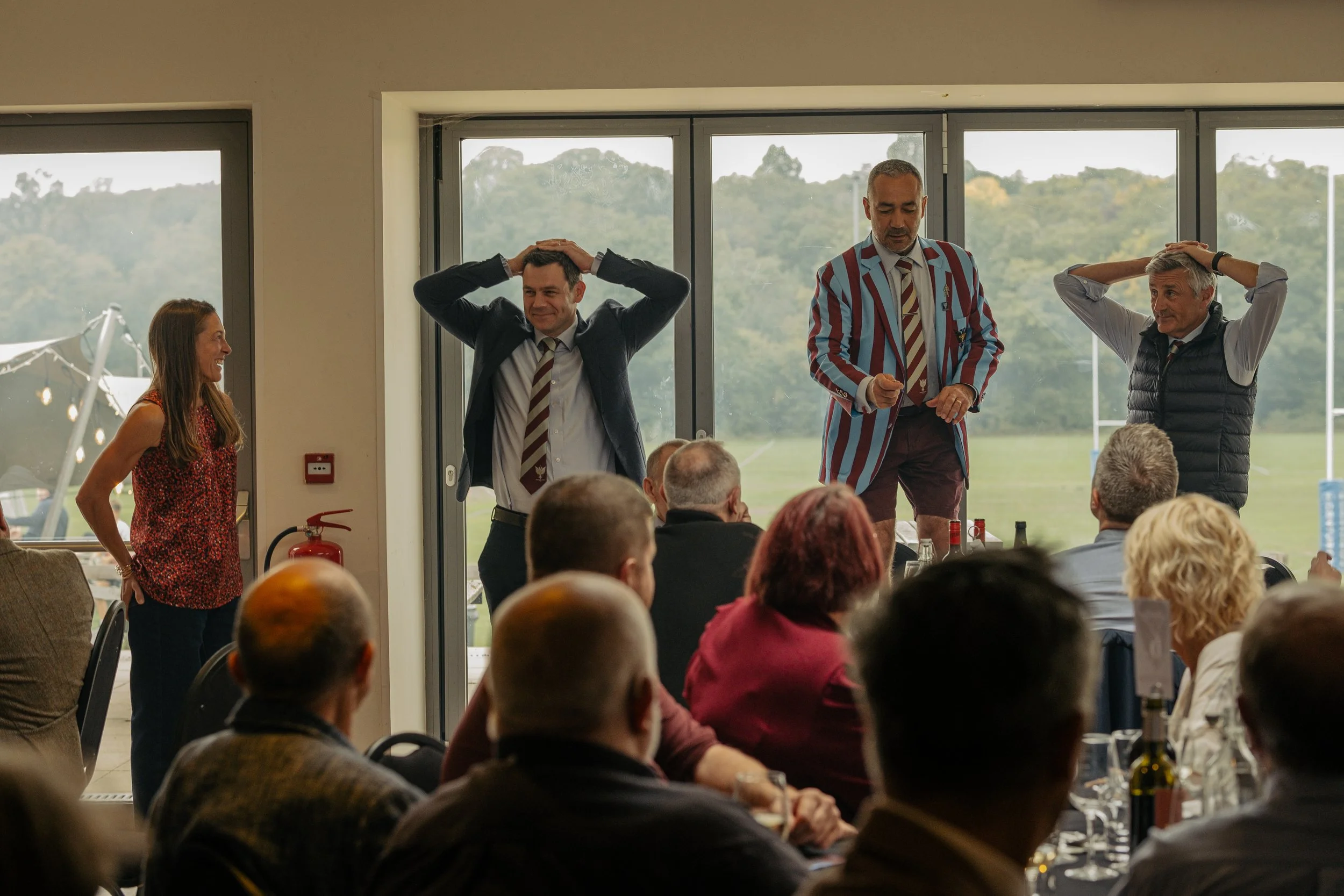 A group of people performing comedy or a skit at an event, with a man in a striped blazer, another in a dark blazer, and an older man on stage, while a woman laughs on the side, in a room with large windows and an outdoor view.