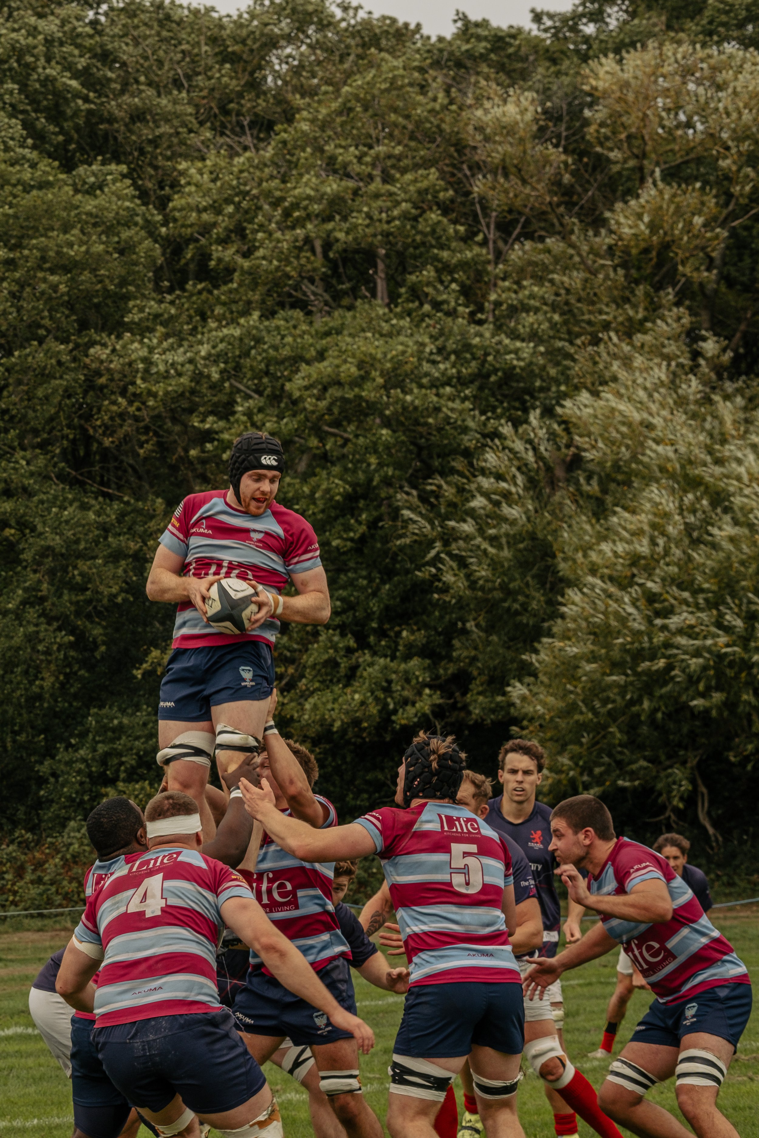 Rugby players in red and gray uniforms lift one player to catch a rugby ball during a game, with others on the field and a background of trees.