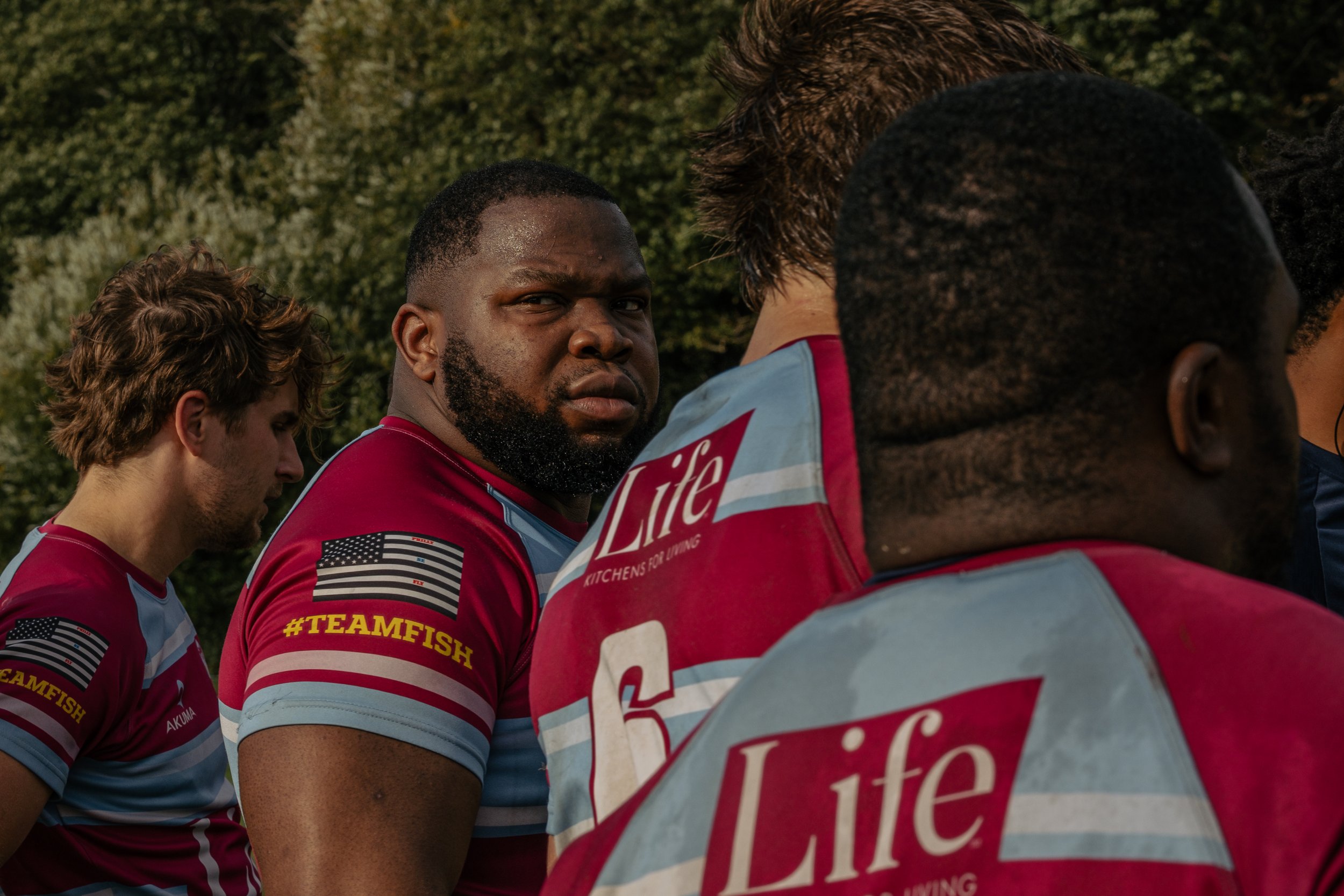 A group of rugby players in maroon and light blue uniforms standing outdoors, with one player looking directly at the camera and others with their backs turned.