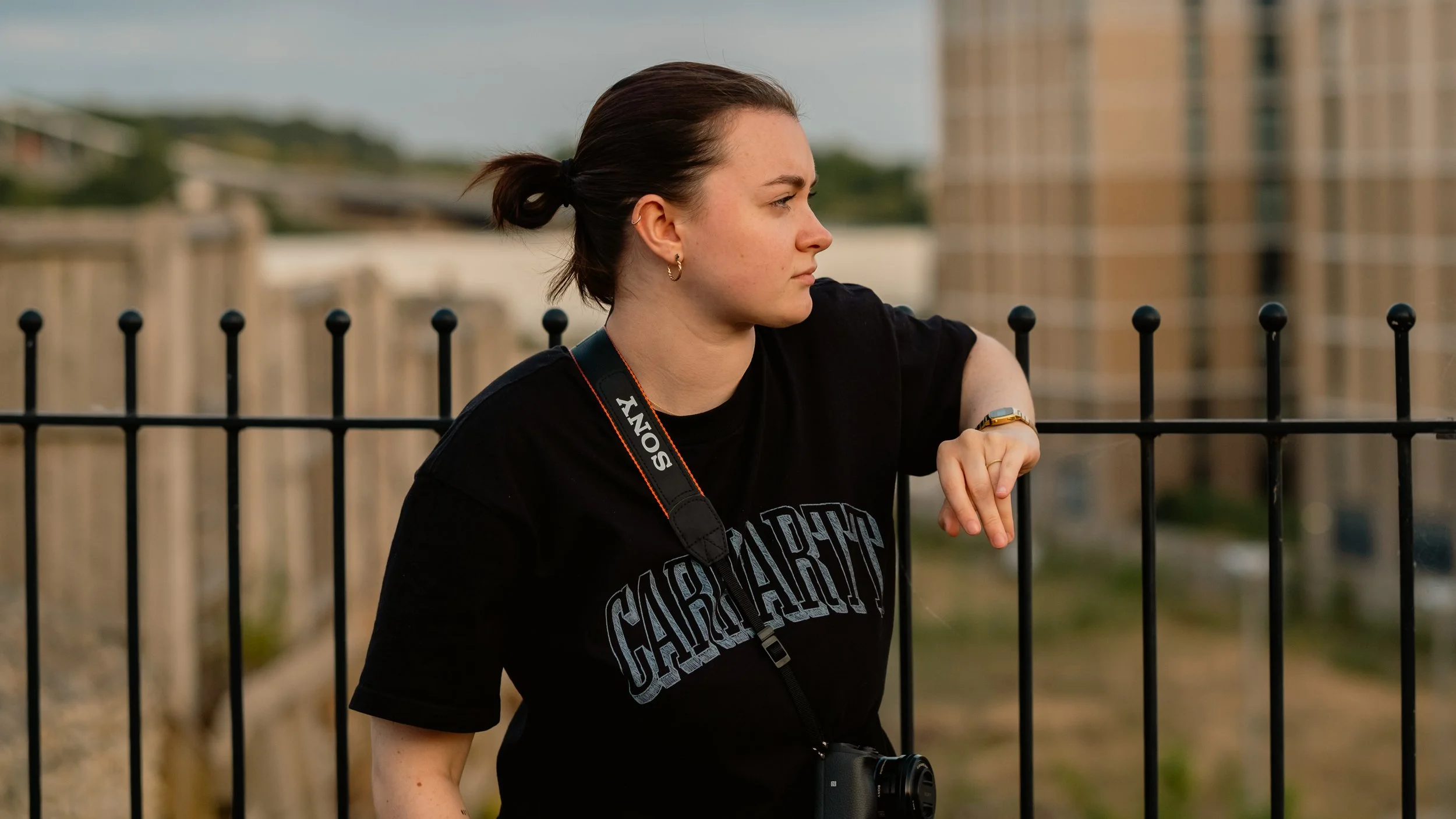 A young woman with dark hair tied in a bun, wearing a black t-shirt with 'Cairn Hart' written on it, standing outdoors near a black metal fence, looking serious or contemplative, with a camera hanging around her neck.
