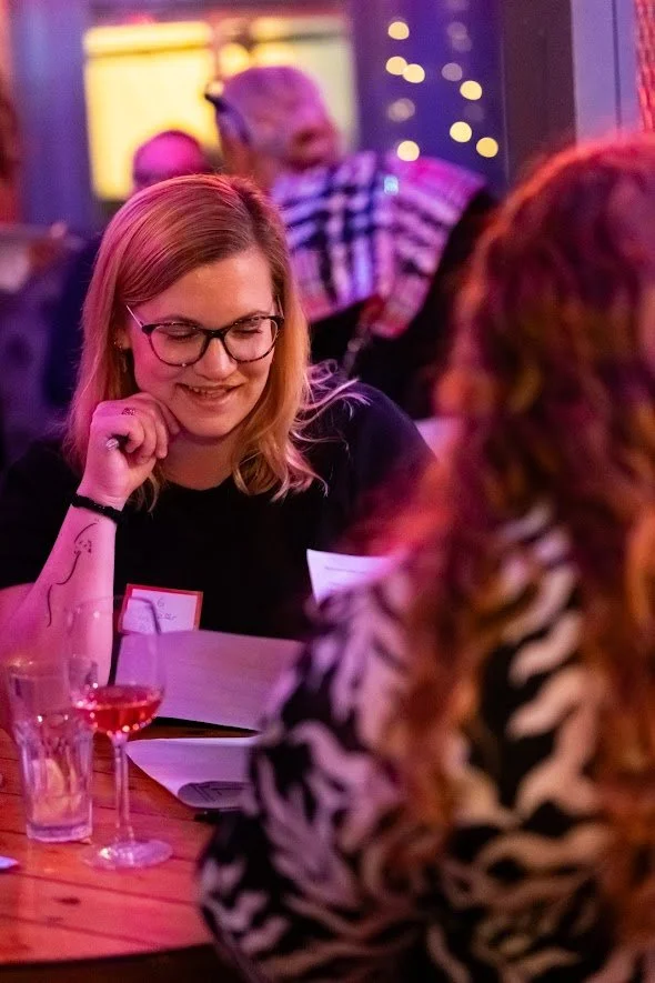 A woman with glasses and blonde hair is sitting at a table, smiling and looking at a person with red hair in a black and white patterned top, in a dimly lit, colorful venue with string lights in the background.