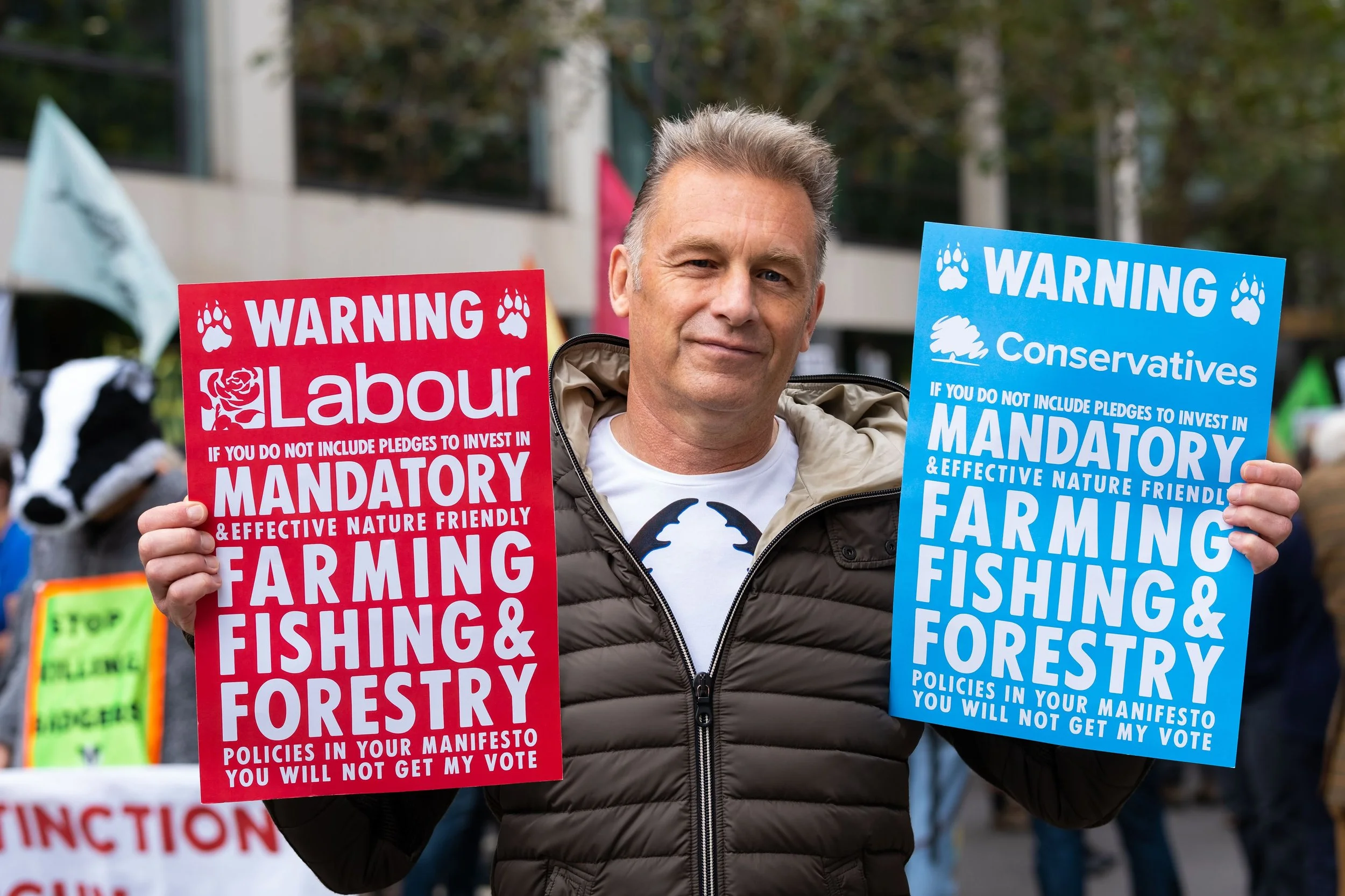 A man holding two protest signs, one red and one blue, during a demonstration. The red sign advocates for mandatory and effective nature-friendly farming, fishing, and forestry policies, while the blue sign criticizes the Conservative party for not i