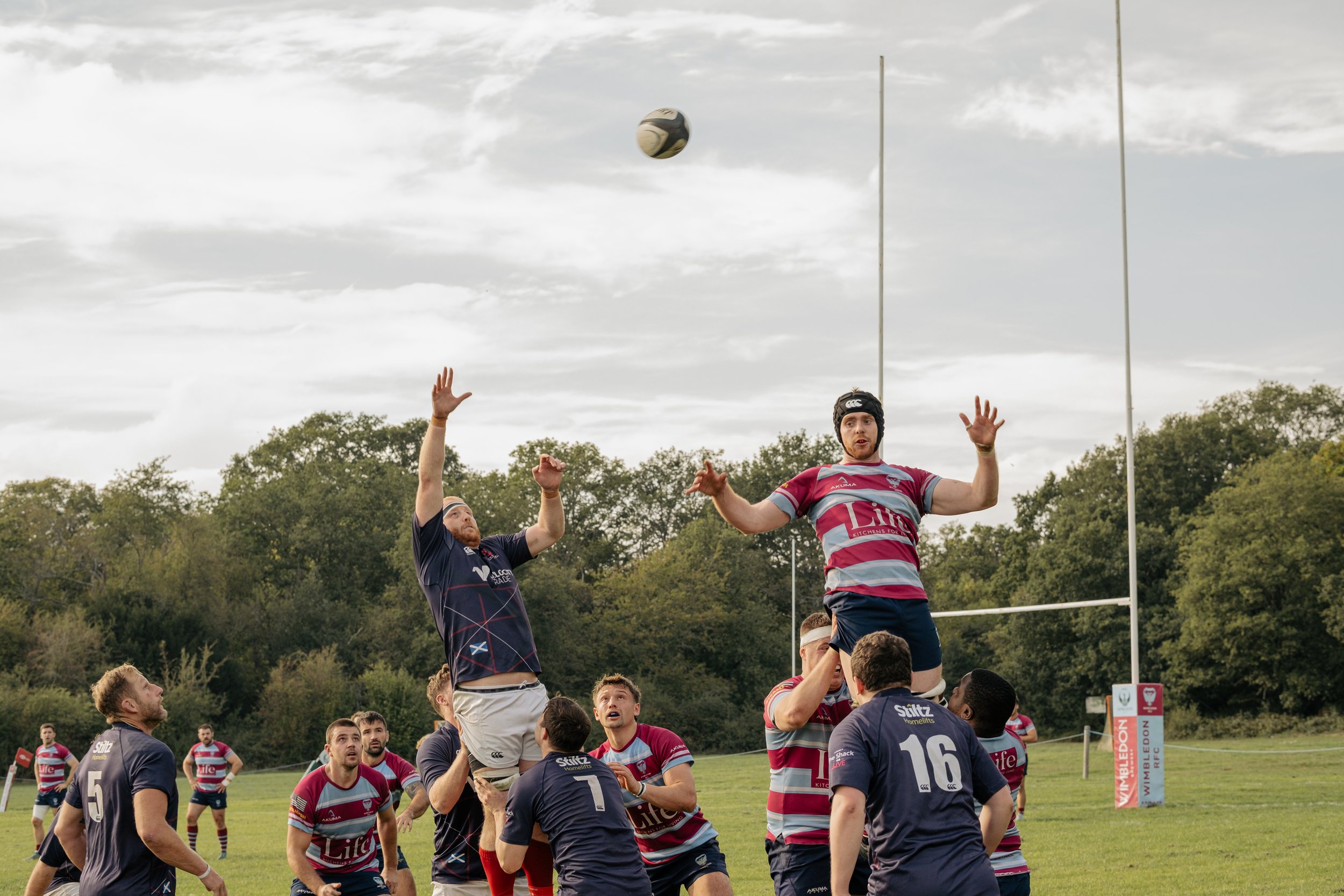 Rugby players in mid-game contest, jumping to catch the ball near the goalposts on a grassy field with trees in the background.