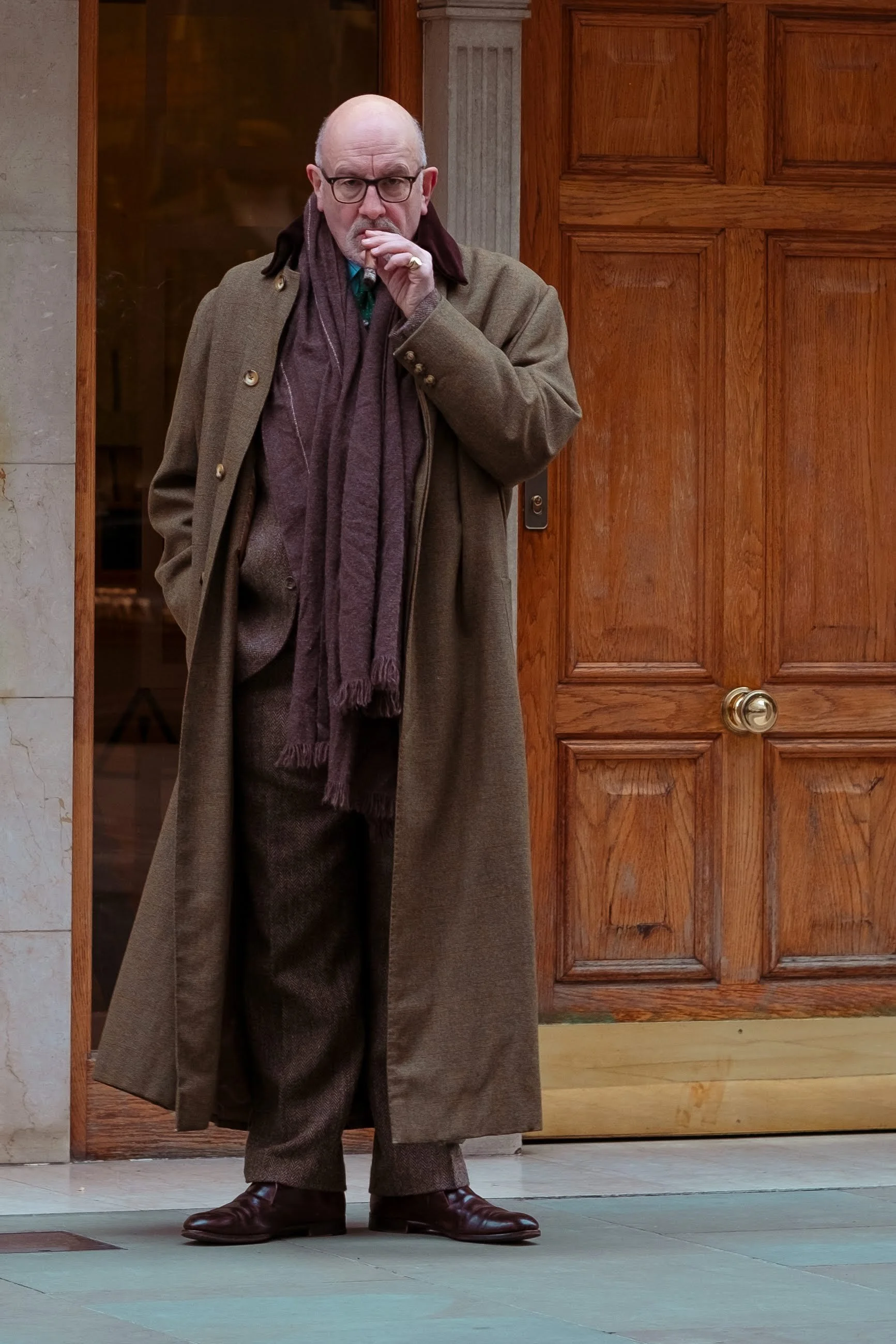 A bald man wearing glasses, a brown coat, and a purple scarf standing outside a wooden door, with his hand near his mouth.