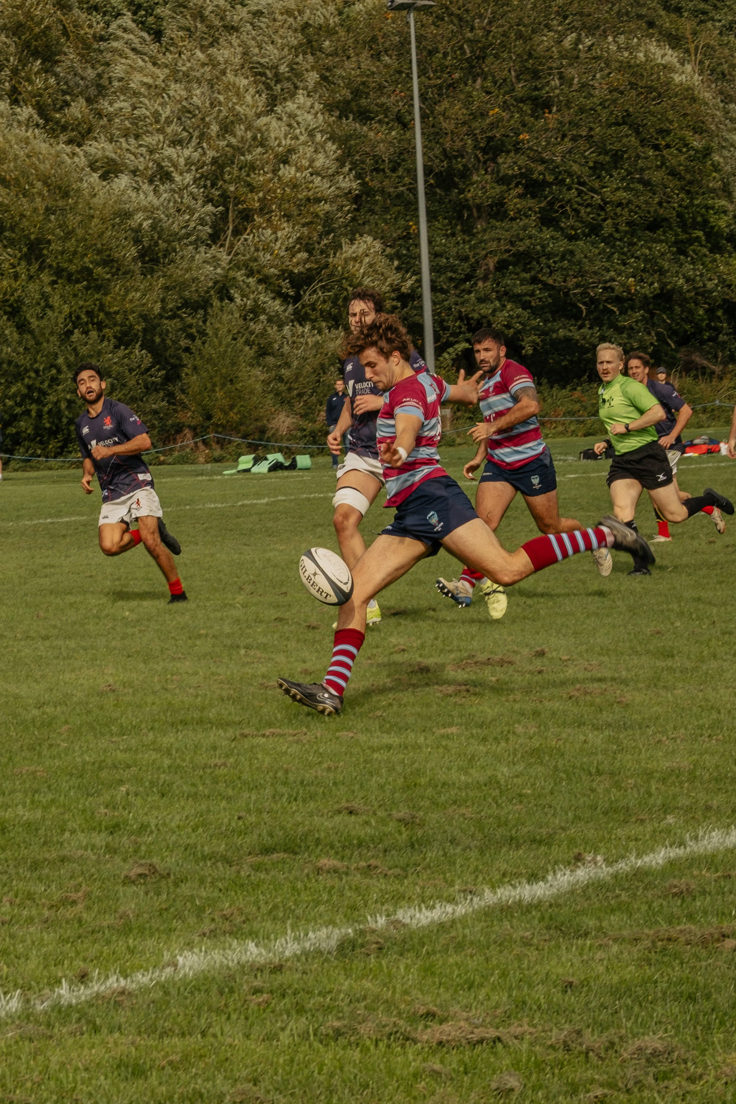 Rugby players in action on a grassy field, with one player kicking the ball while others chase.