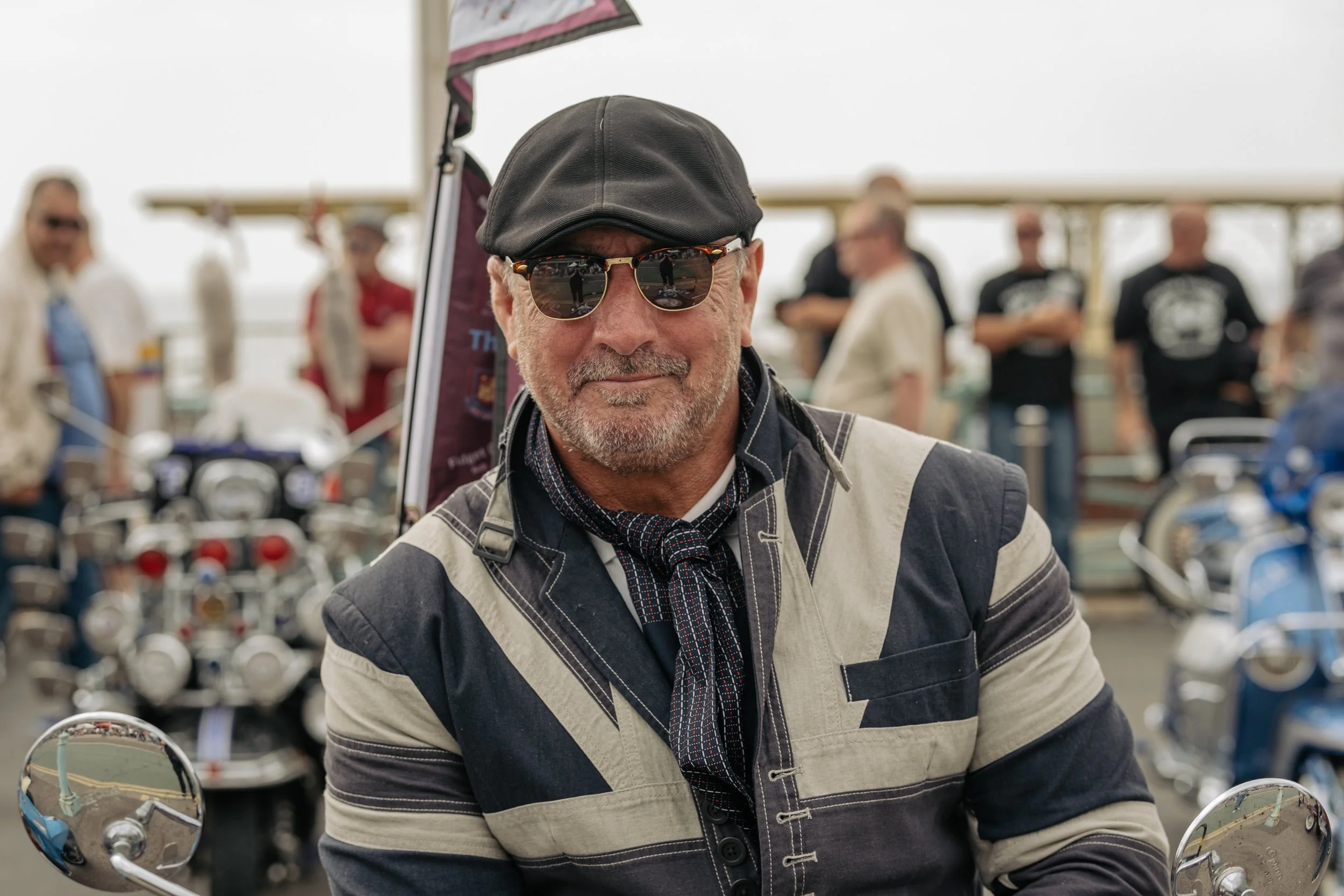 A man with a gray beard wearing sunglasses, a black cap, a scarf, and a patterned jacket, sitting on a motorcycle at an outdoor event with a group of people and motorcycles in the background.