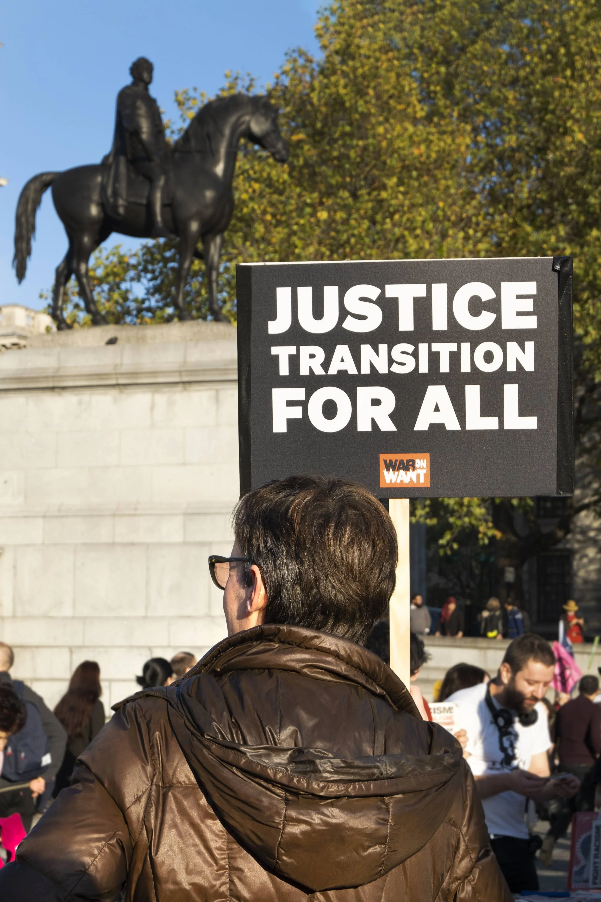 A person holding a protest sign that reads 'Justice Transition for All' during a protest or demonstration, with an equestrian statue in the background and other people gathered around.