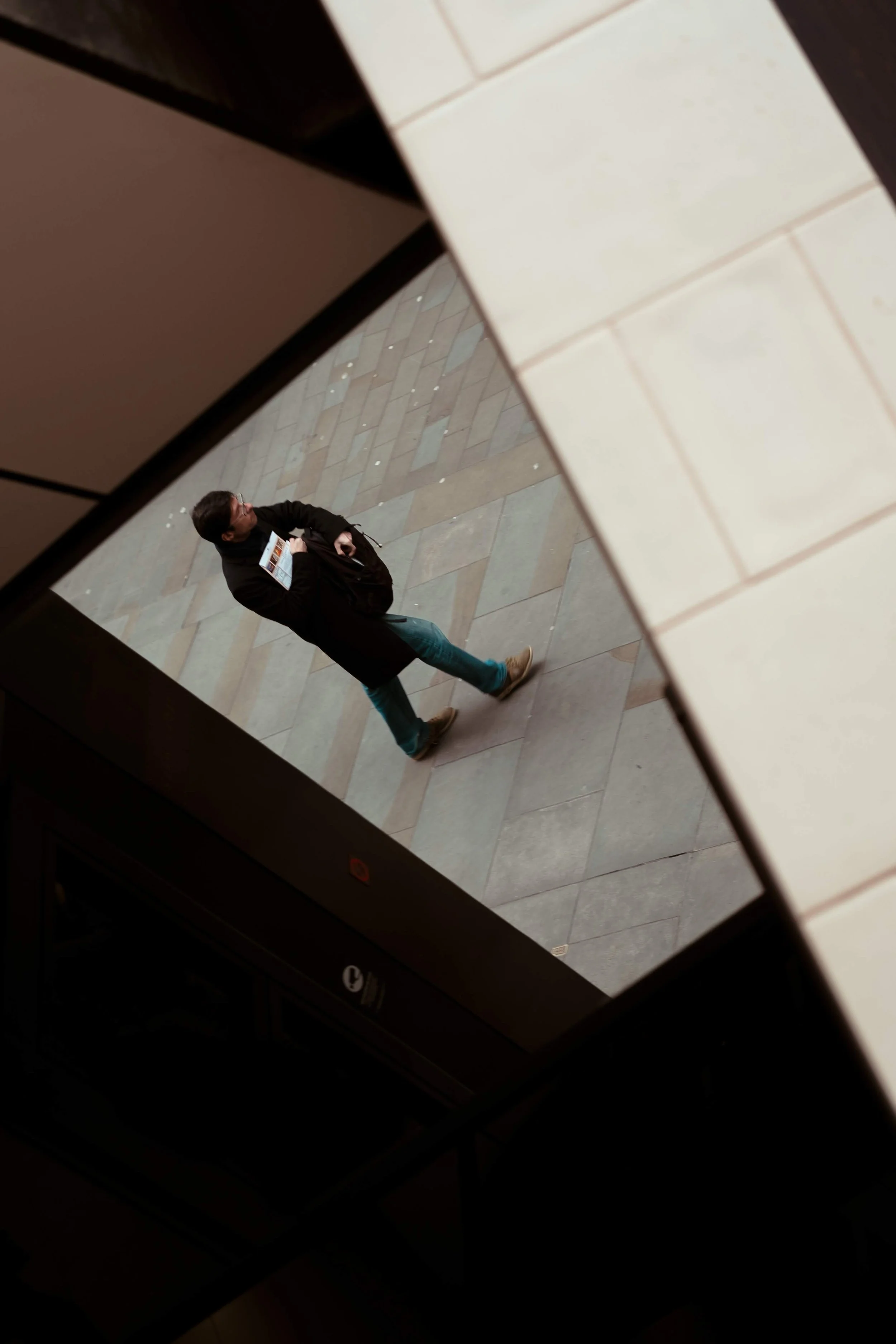 A man in a black jacket and teal jeans walking on a tiled sidewalk viewed from above through a window.