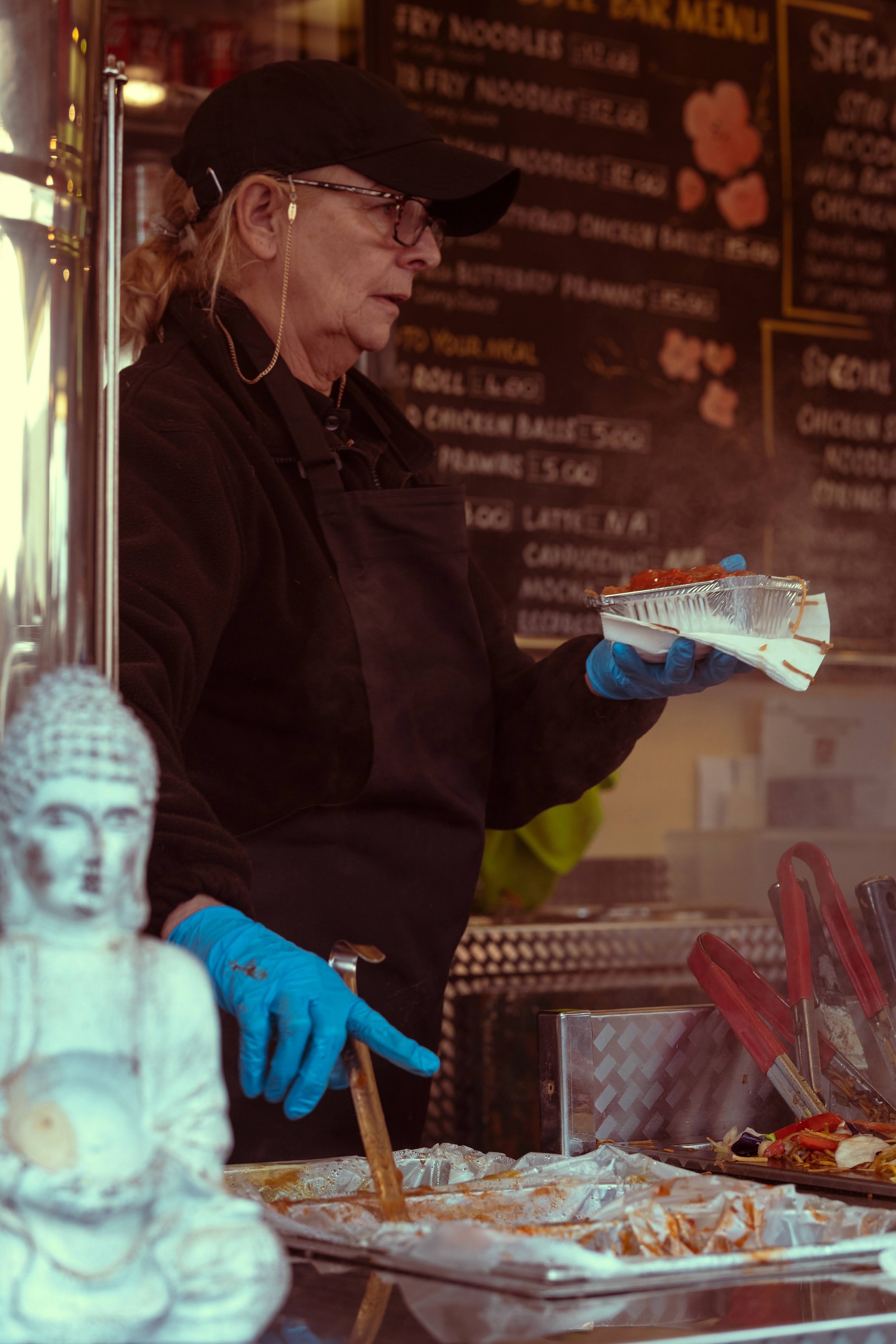 A woman wearing glasses, a black cap, and blue gloves prepares food at a food stall. She is holding a paper tray with food in one hand and stirring with a utensil in the other. There is a blackboard with a menu in the background and a decorative Budd