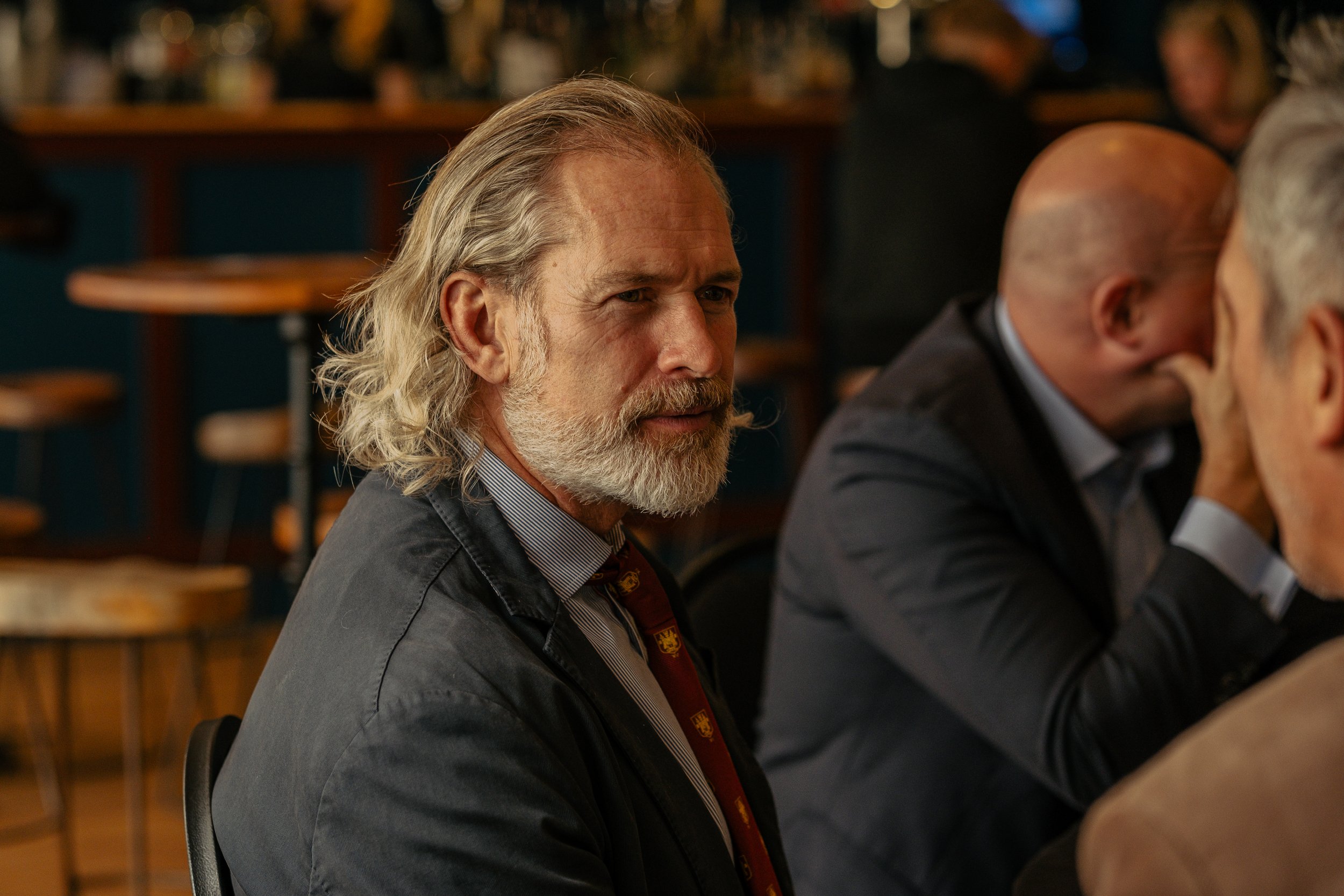 A man with gray hair and a beard, dressed in a suit, seated at a table in a social or business setting.